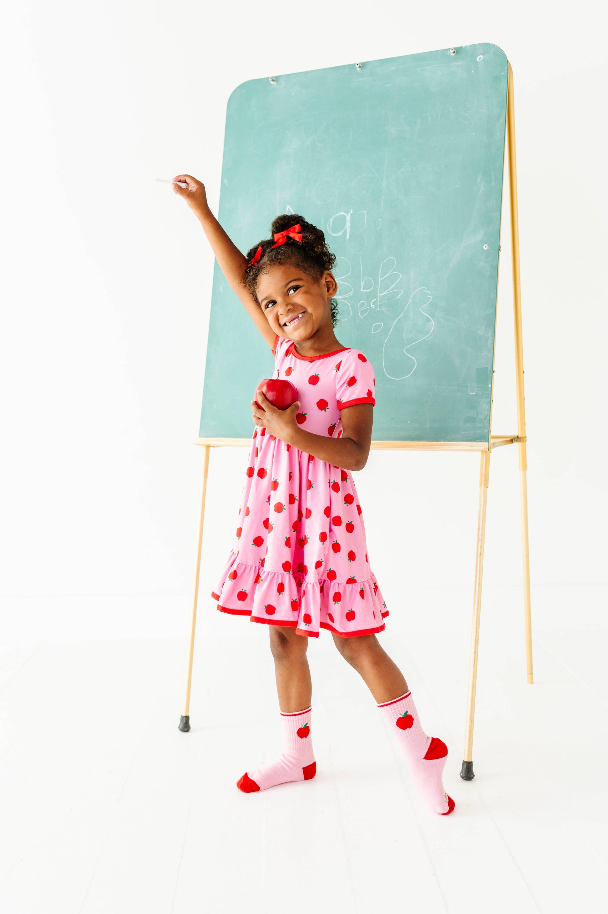 A young girl with curly hair wearing a pink and red polka-dot dress and heart-patterned socks stands in front of a green chalkboard easel, holding a red apple and raising her arm in a playful classroom-themed pose.