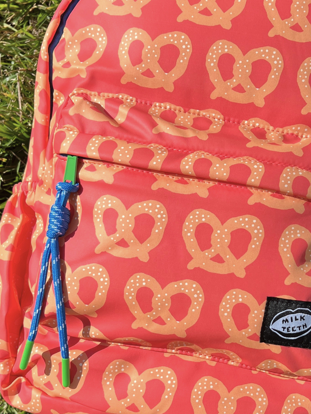 A close-up of a red backpack with a repeating pretzel pattern, featuring a blue and green drawstring and a black 'Milk Teeth' brand patch.