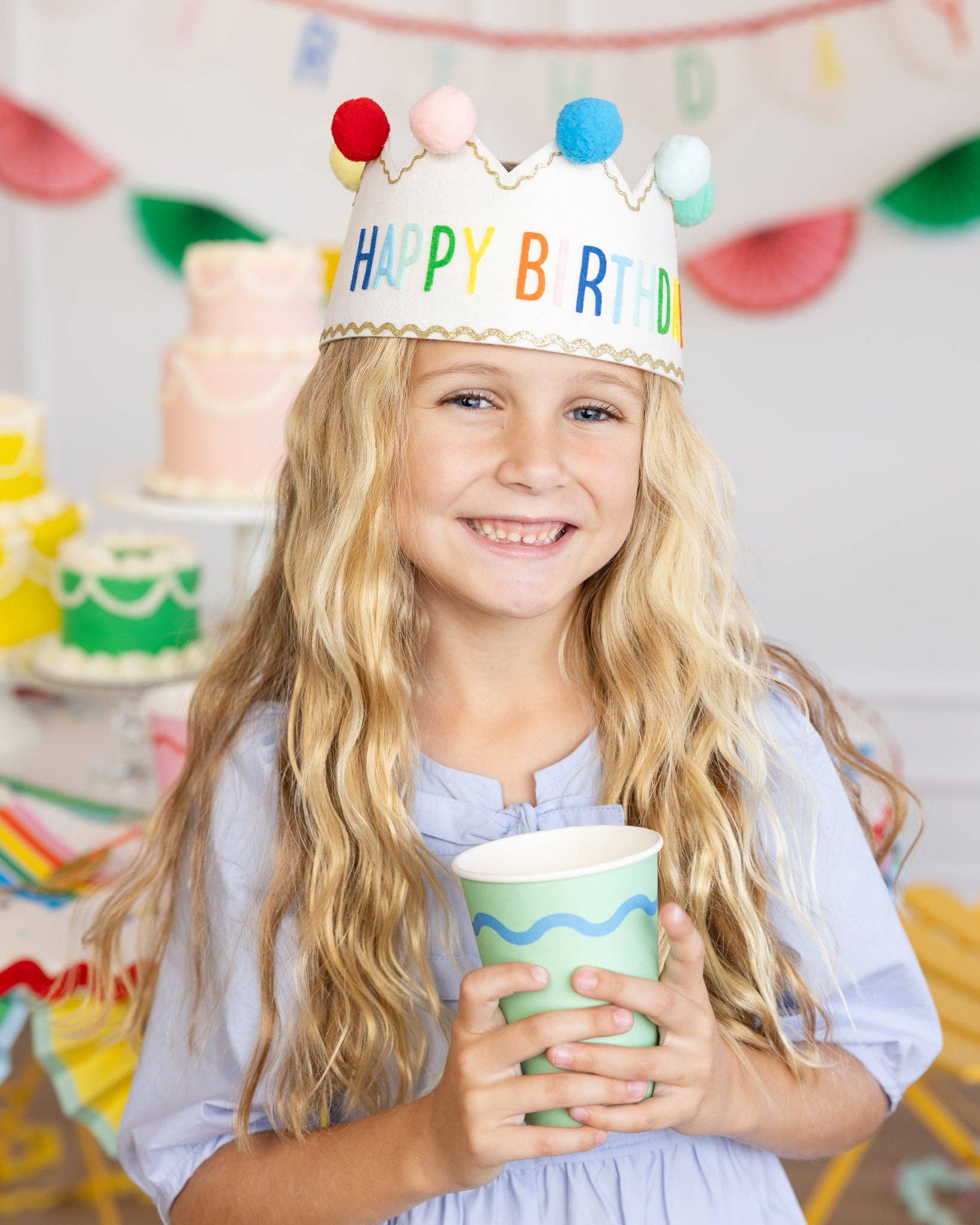 Young girl wearing a colorful 'Happy Birthday' crown, holding a paper cup, with a festive background.