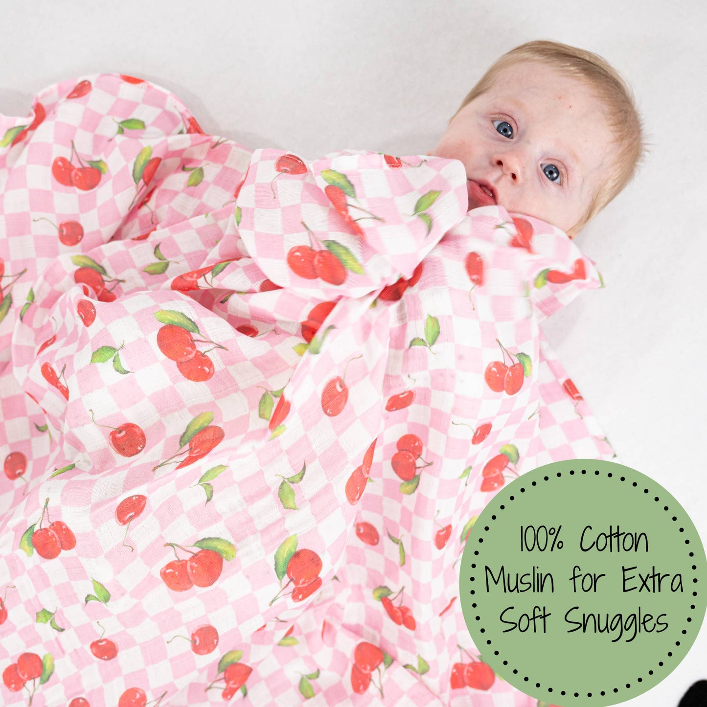Baby wrapped in a pink cherry-patterned blanket with a green label indicating 100% cotton muslin.