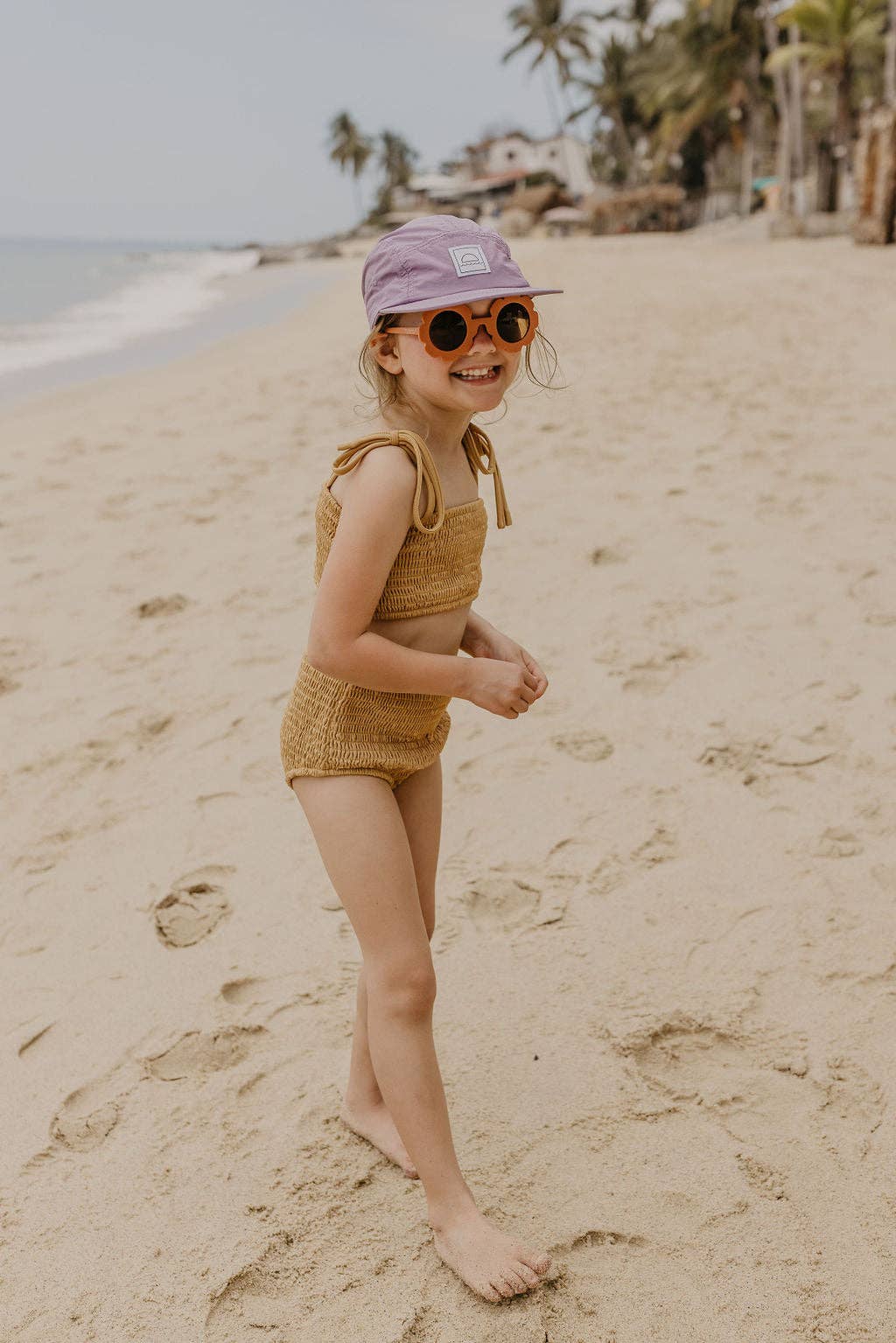 Child in a yellow swimsuit and purple hat standing on a sandy beach with palm trees in the background.