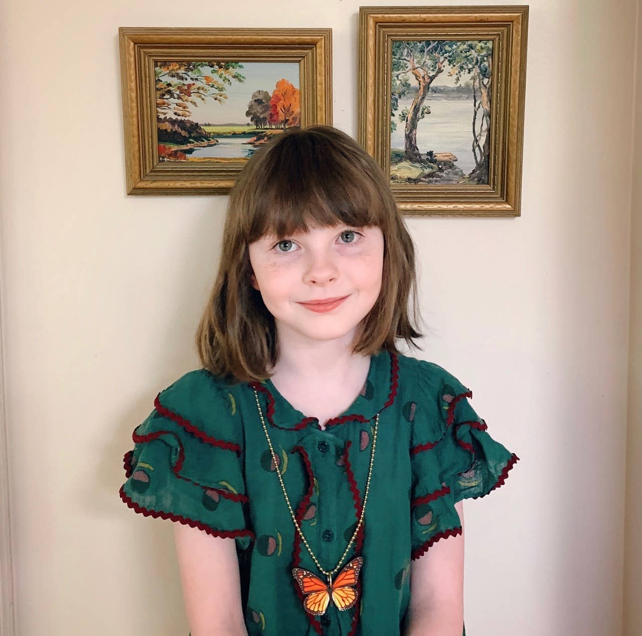 Young girl wearing a green dress with butterfly design in front of two framed paintings.