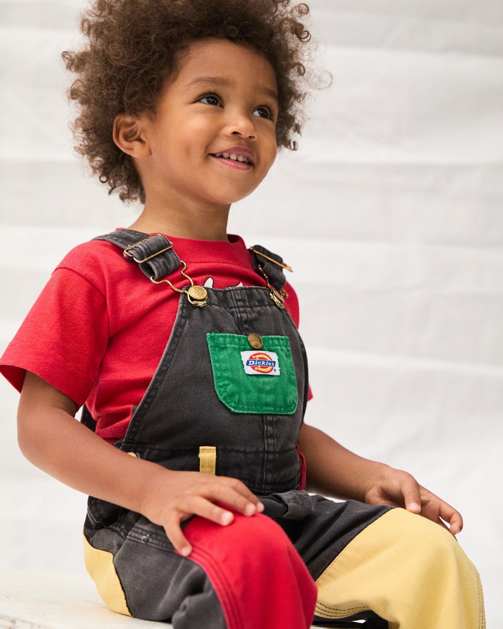 Child wearing a red shirt and black overalls with a green pocket patch on a white background