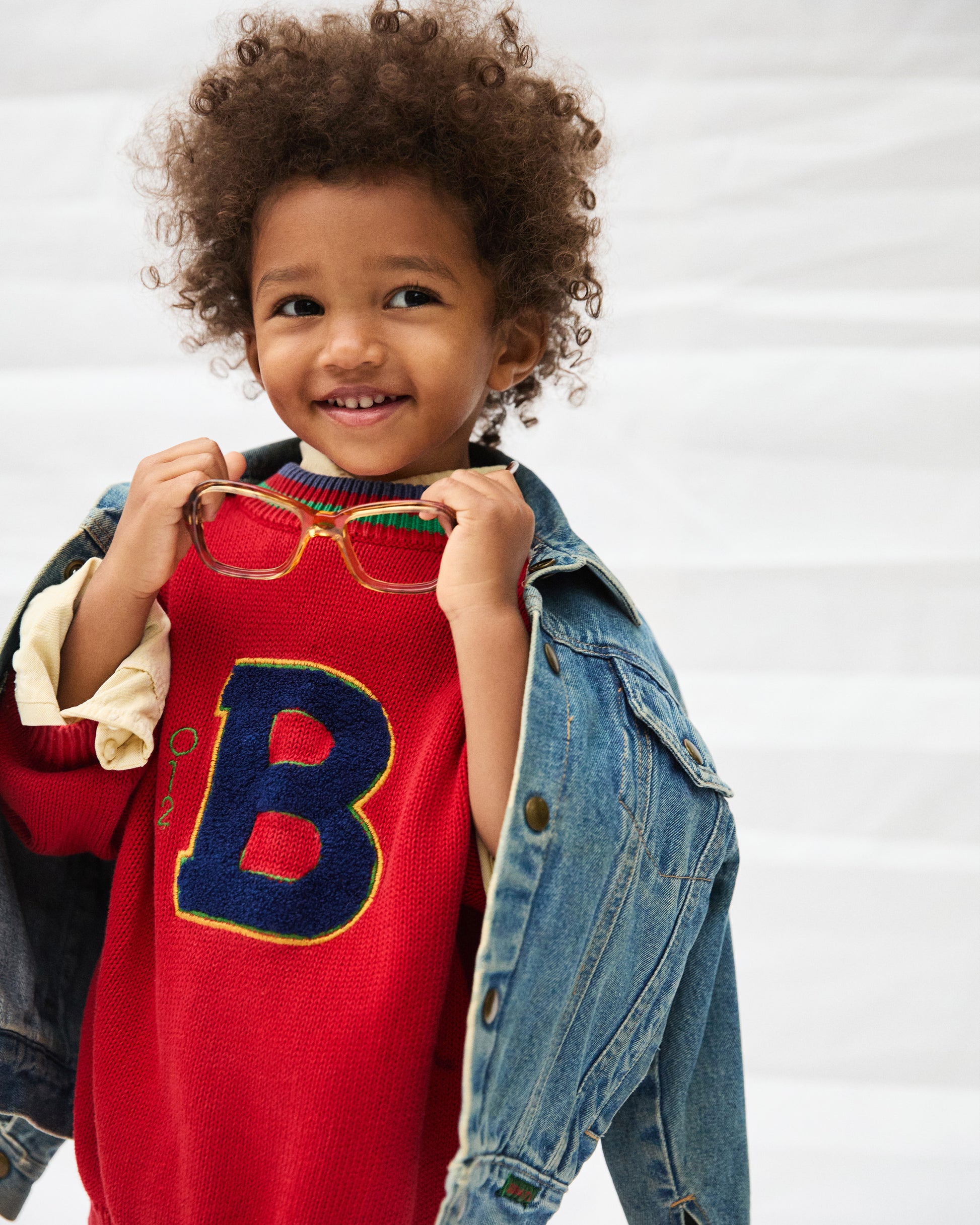 Child wearing a red sweater with a large letter 'B' and holding glasses, standing against a plain background.