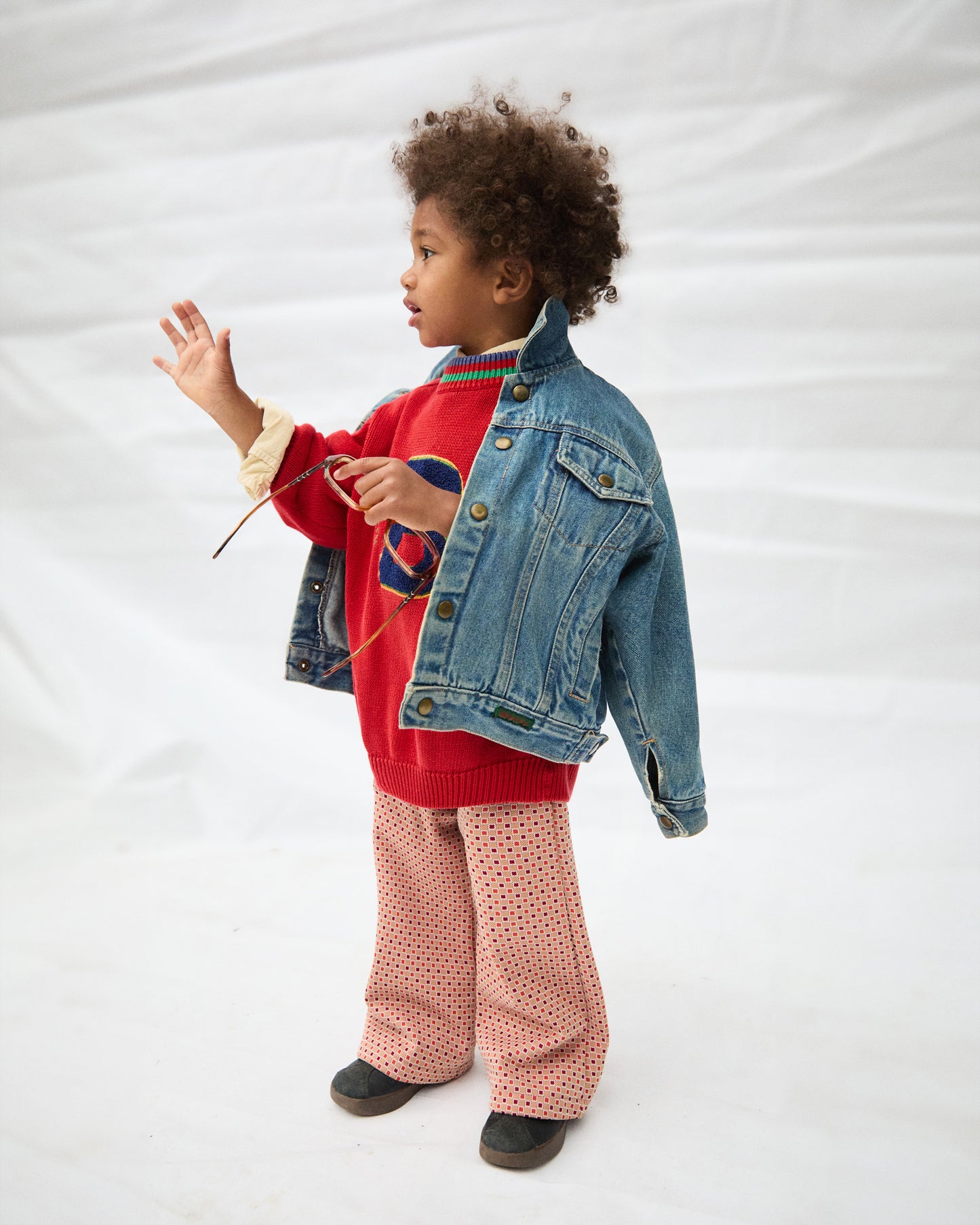 Child wearing a red sweater, denim jacket, and checkeredpants on a white background