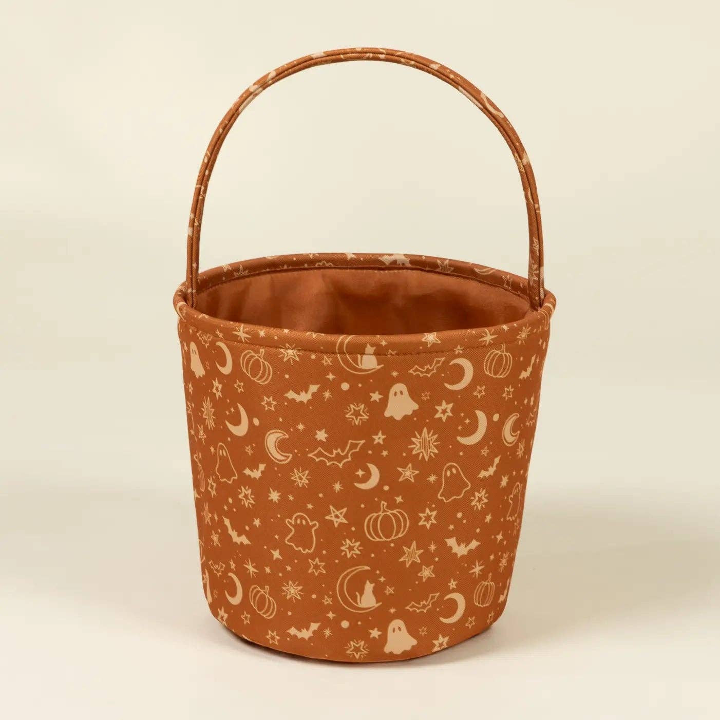 A small fabric bucket with a rounded top handle, featuring a rust-orange body printed with white Halloween motifs like moons, ghosts, pumpkins, and stars, set against a plain cream background.