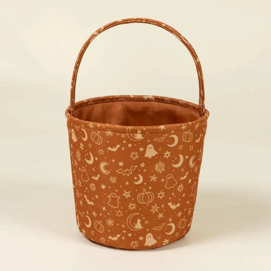 A small fabric bucket with a rounded top handle, featuring a rust-orange body printed with white Halloween motifs like moons, ghosts, pumpkins, and stars, set against a plain cream background.
