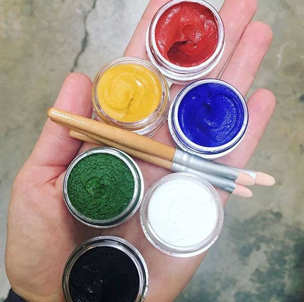 Hand holding six small containers of colored paint with corresponding paintbrushes.
