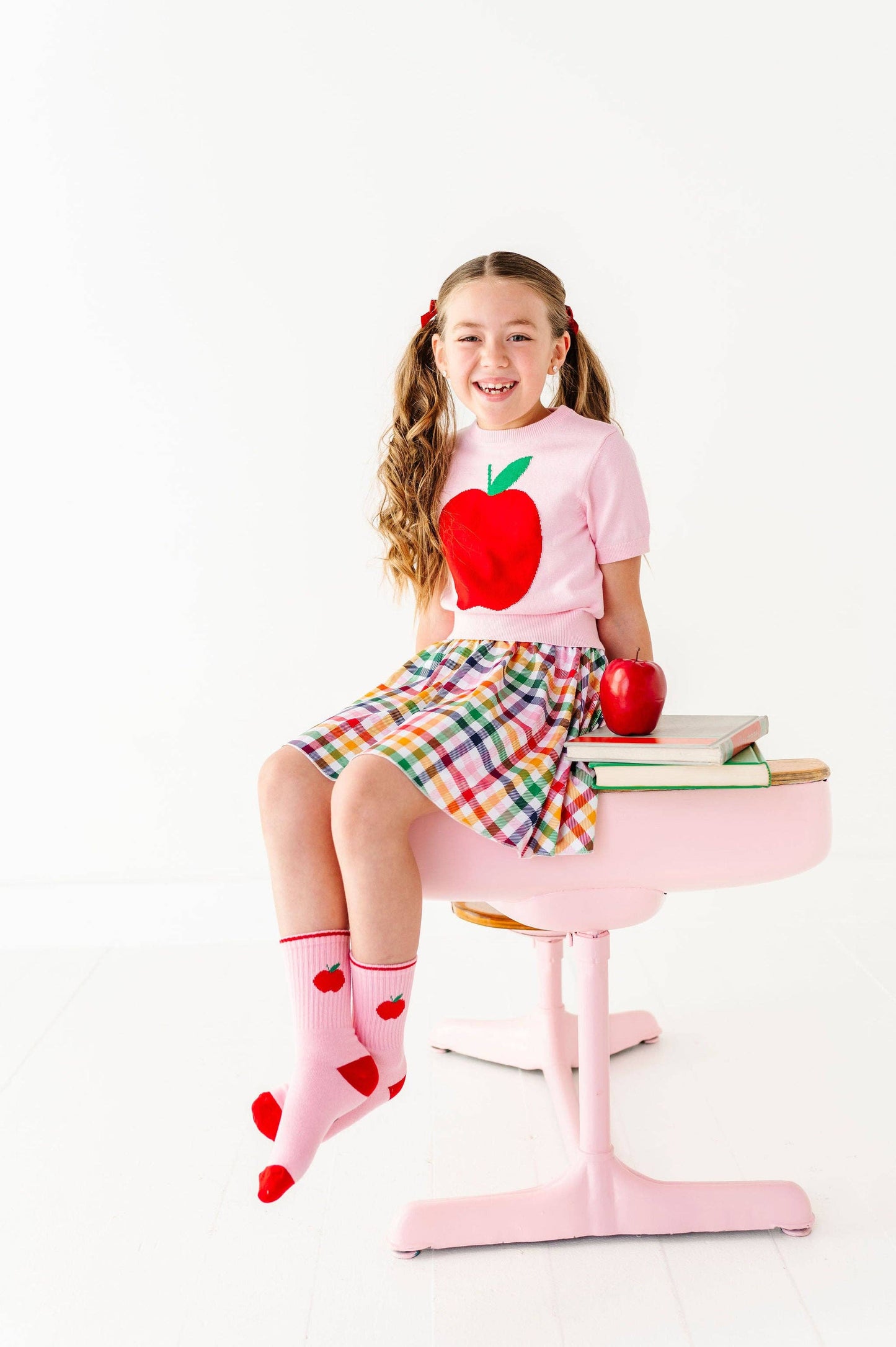 A young girl with pigtails sits on a pink school desk wearing a pink t-shirt with a red apple graphic, a plaid skirt, and pink socks with red hearts. A red apple and books are on the desk against a white studio background.