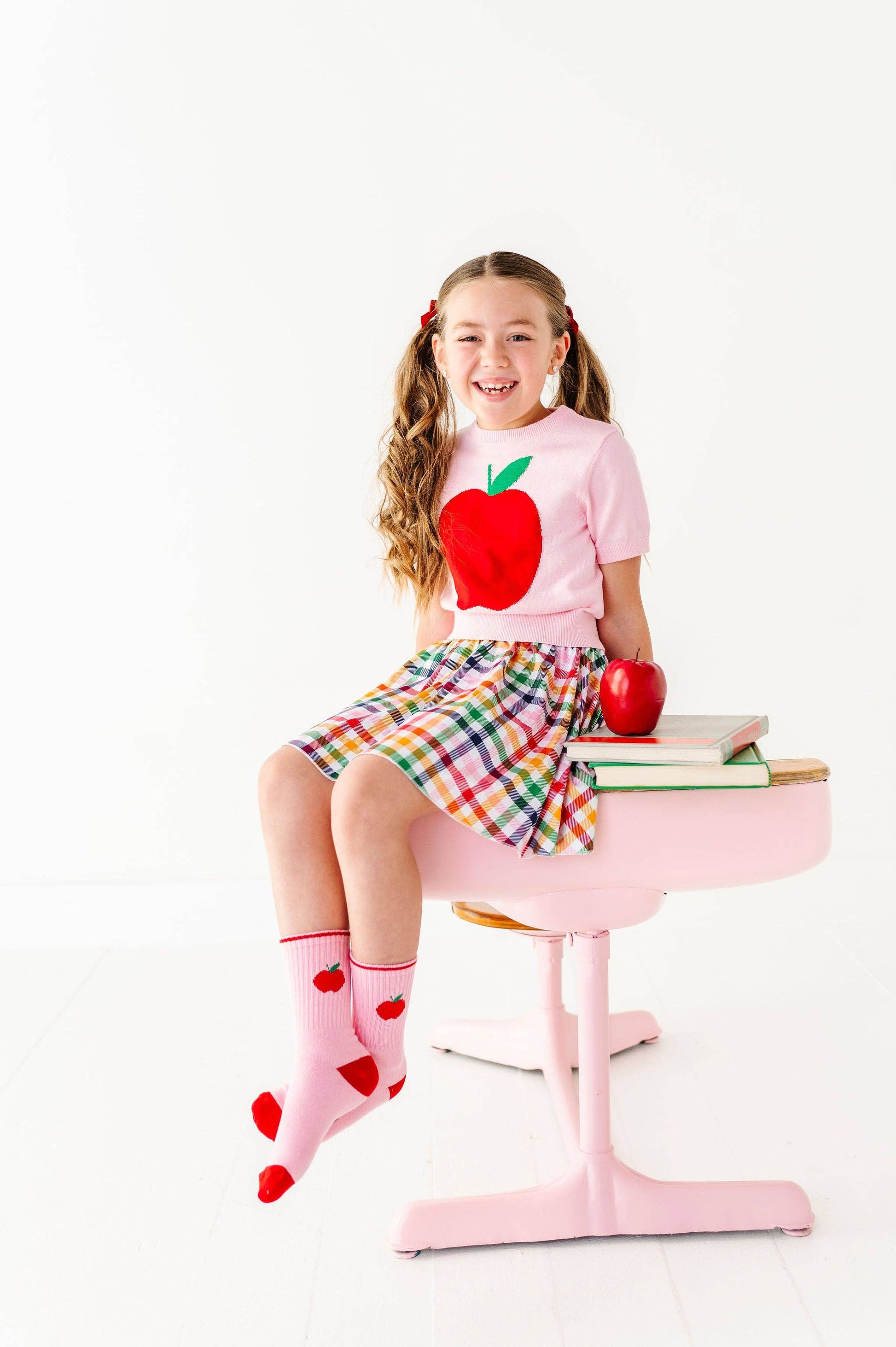 A young girl with pigtails sits on a pink school desk wearing a pink t-shirt with a red apple graphic, a plaid skirt, and pink socks with red hearts. A red apple and books are on the desk against a white studio background.