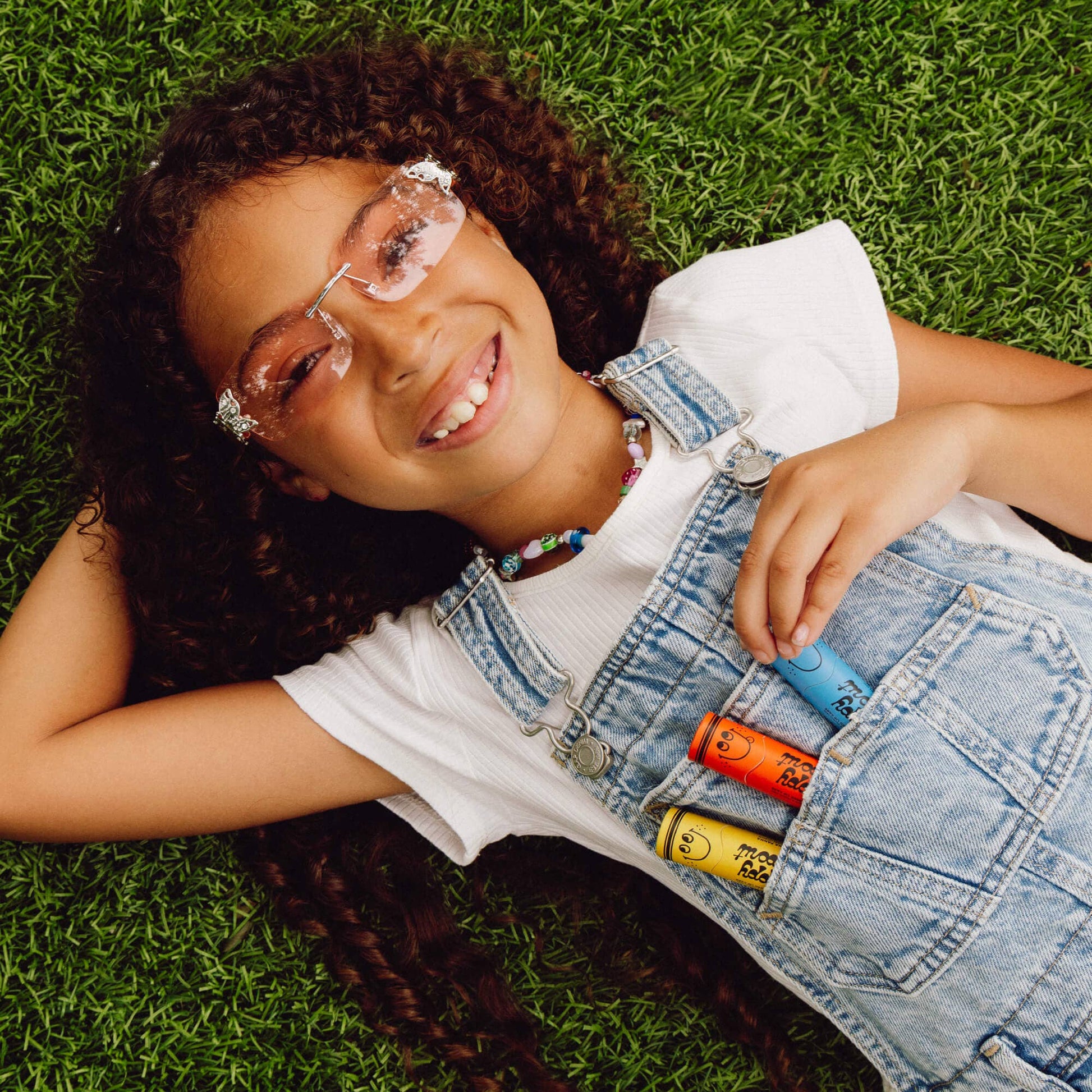 Child wearing glasses and denim overalls lying on grass with colorful lip balm.