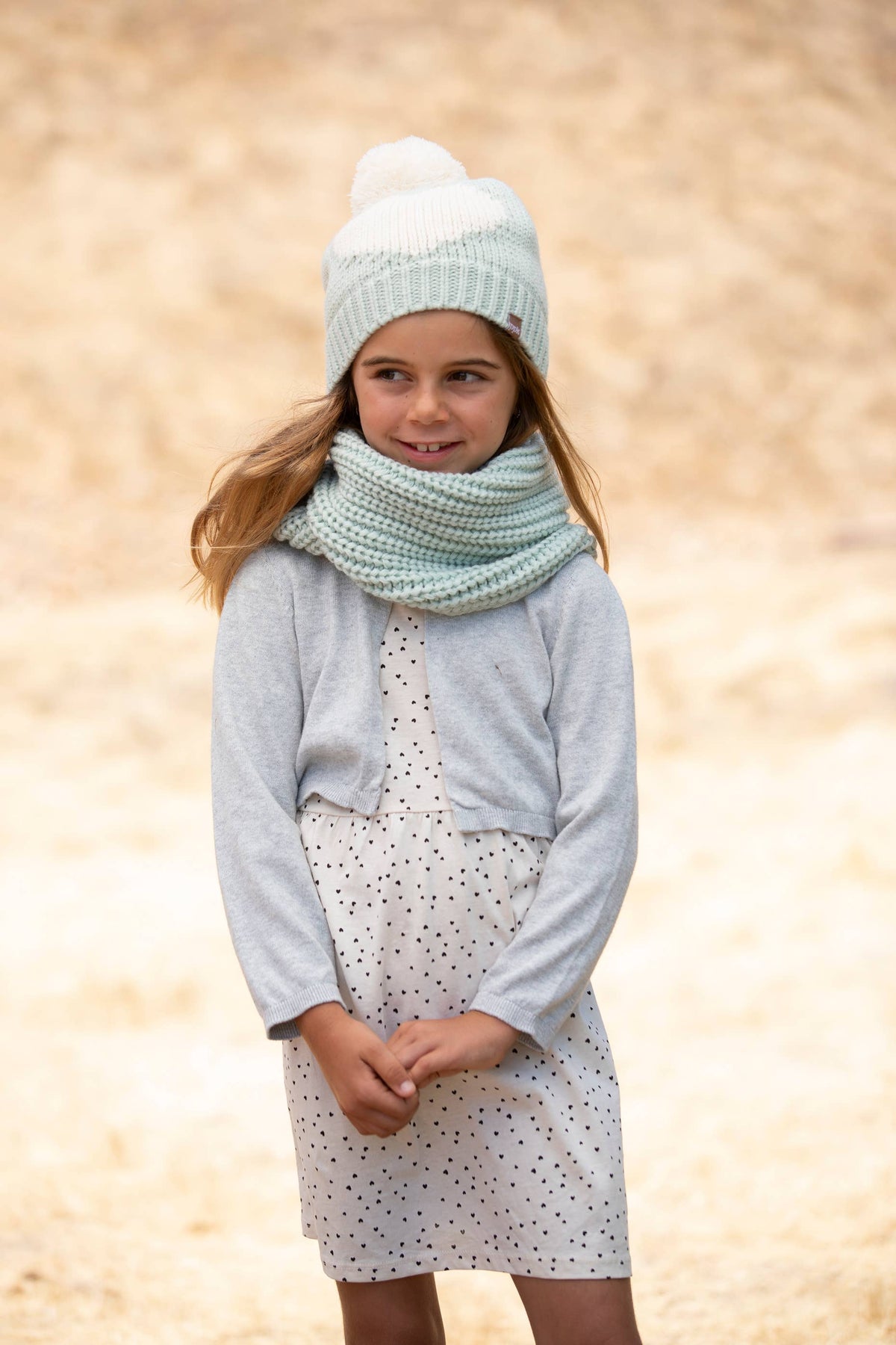 A young girl wearing a light blue knit beanie with a pom-pom, a chunky light blue knit scarf, a light gray cardigan, and a white dress with small black polka dots, standing against a warm sandy background.