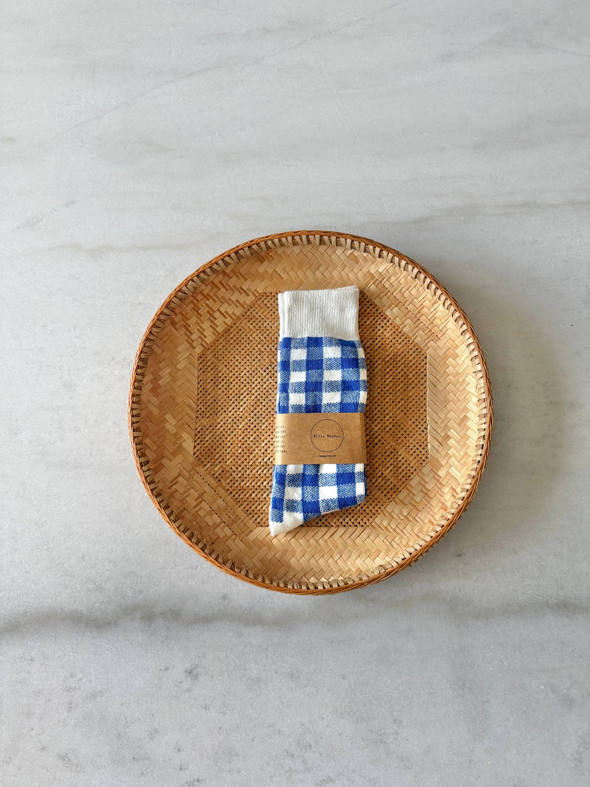 A pair of folded blue and white gingham socks, wrapped in a tan paper band, displayed on a round woven straw plate over a light gray marble surface.