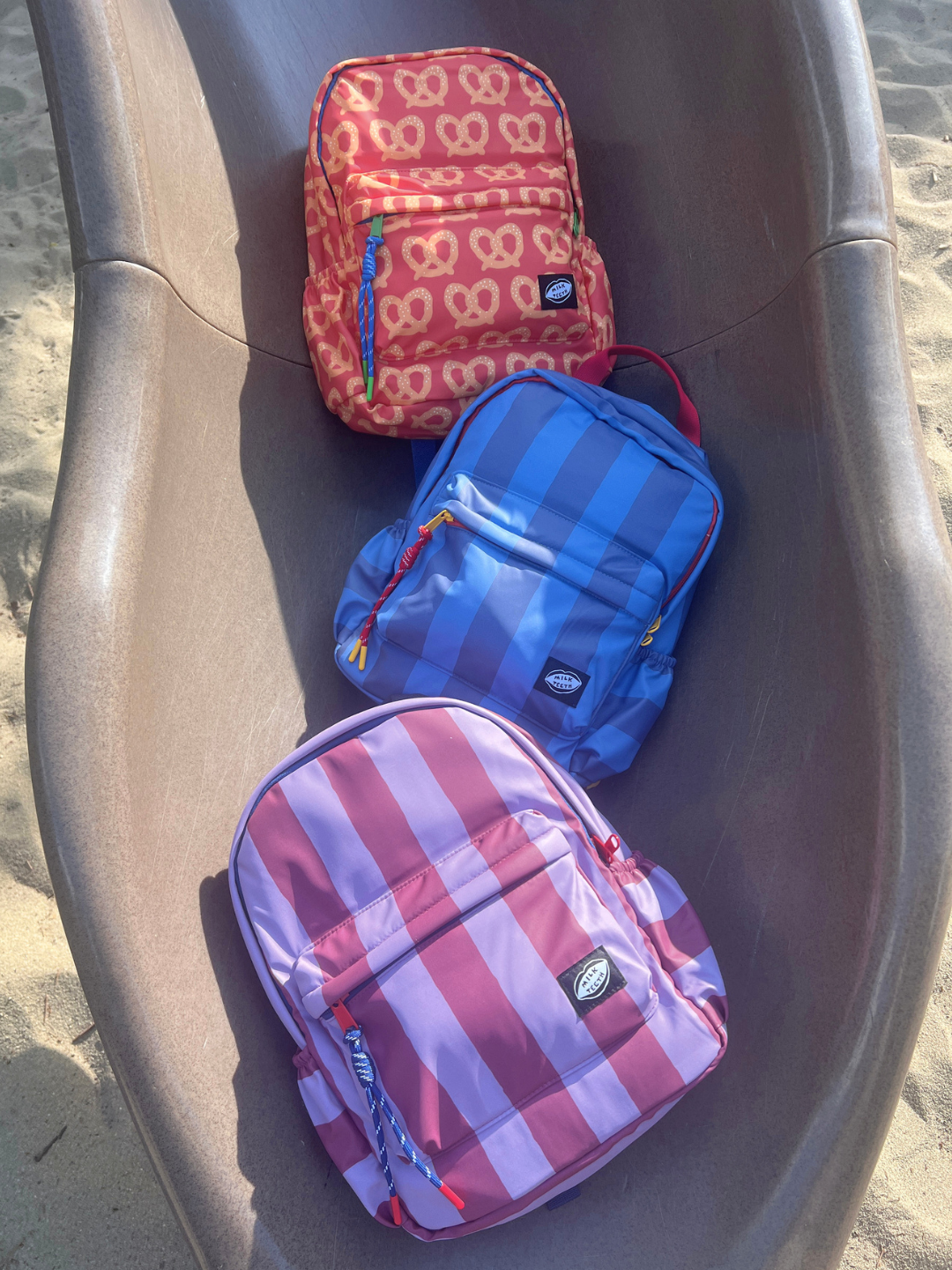 Three backpacks in pink, blue, and red-and-white striped patterns, arranged on a brown plastic slide outdoors on sand.