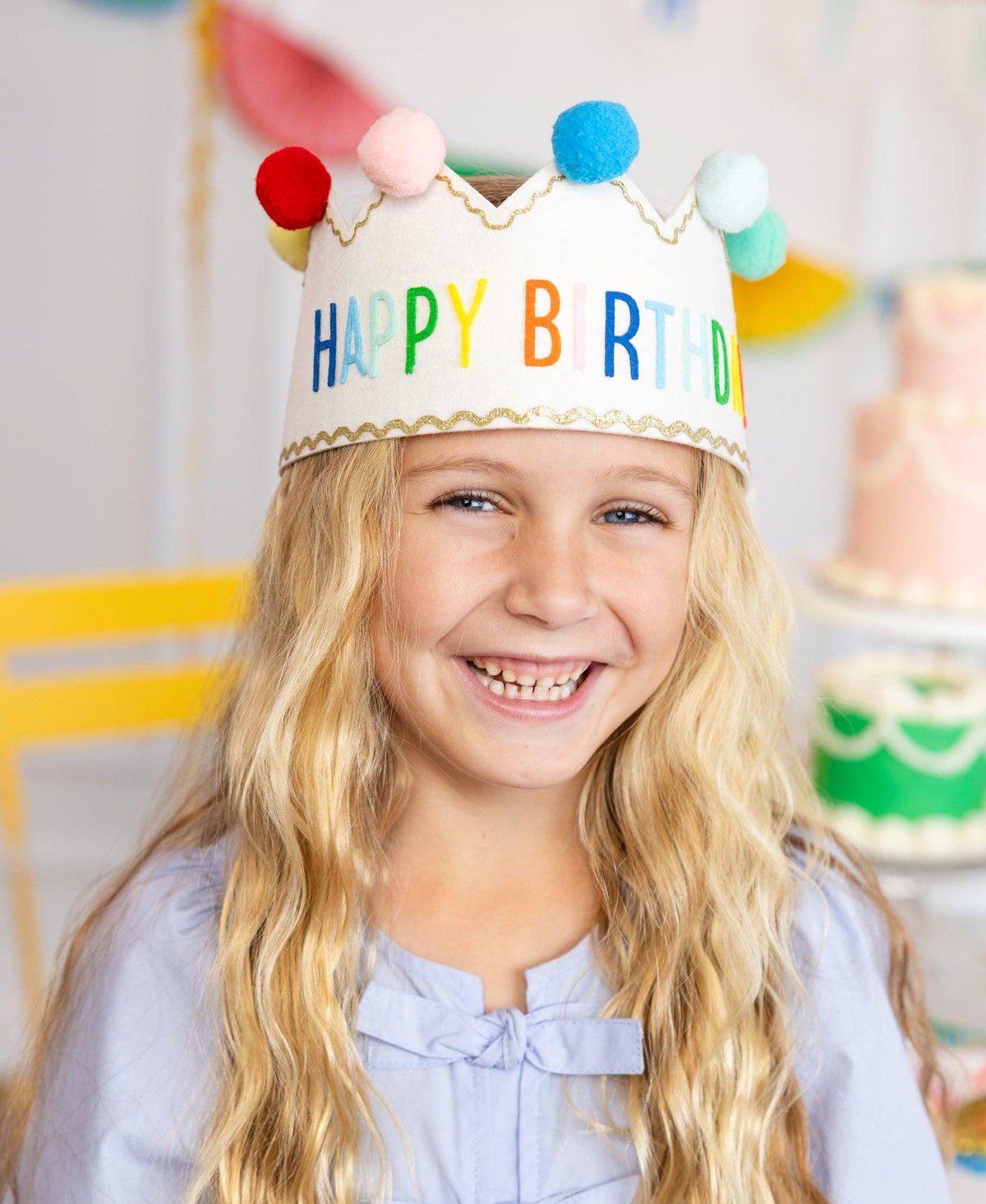 Child wearing a colorful 'Happy Birthday' crown with pom-poms, smiling in a festive setting.