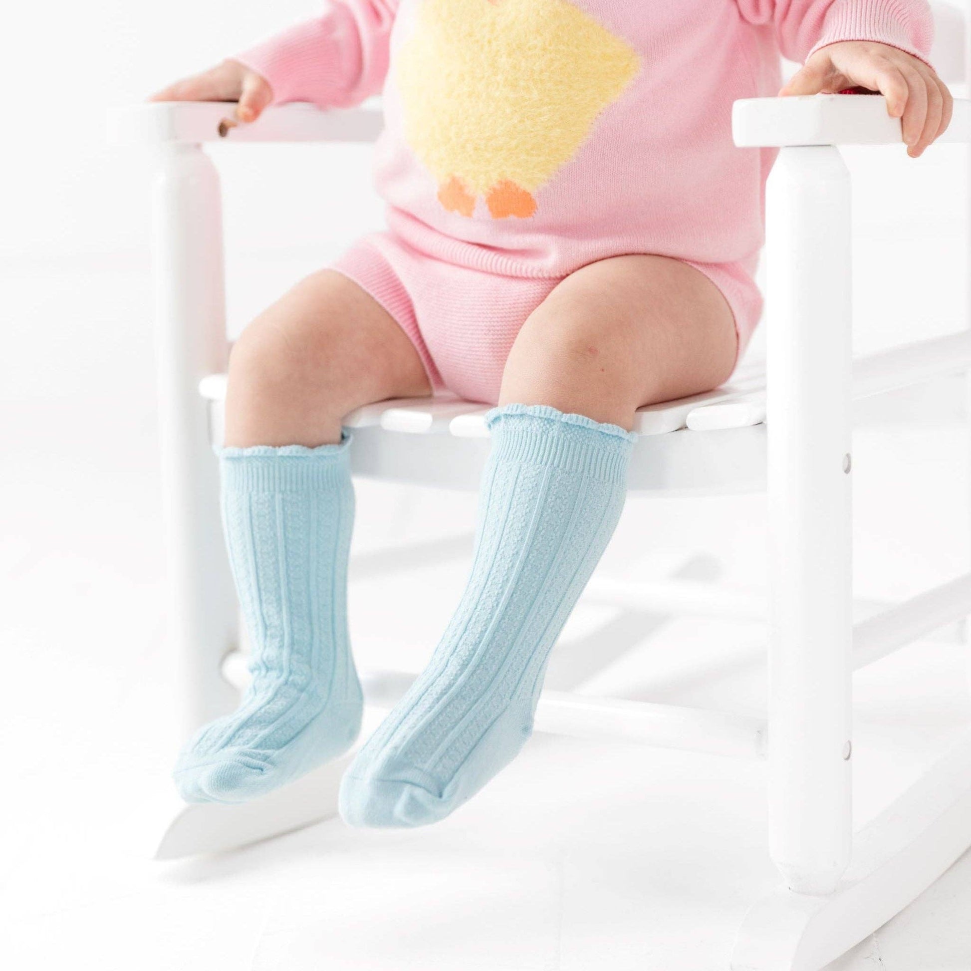 Baby wearing light blue socks and a pink outfit sitting on a white chair.