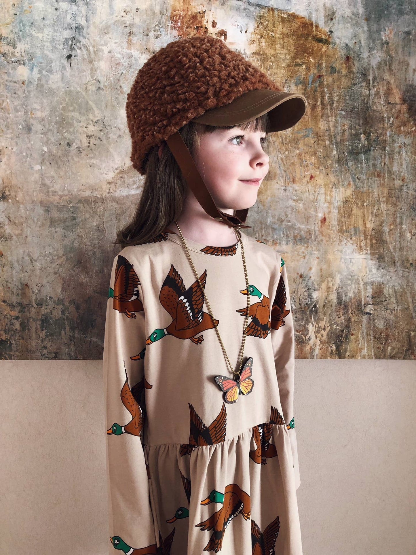 Child wearing a patterned dress and brown hat against a textured wall.