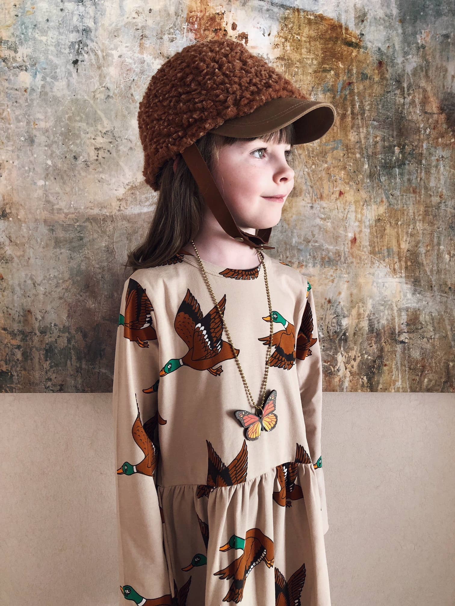 Child wearing a patterned dress and brown hat against a textured wall.