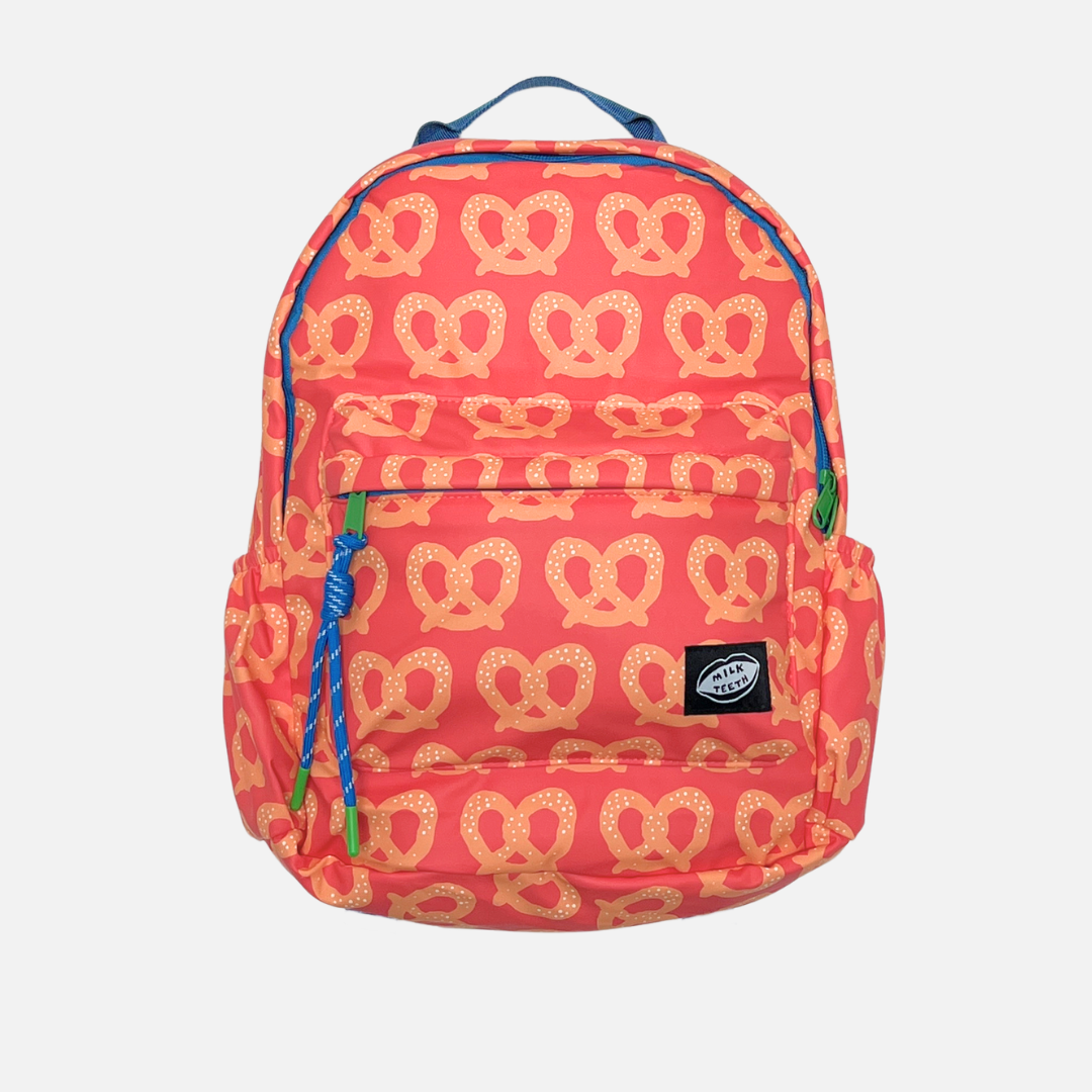 A child's backpack in a coral pink color with an all-over tan pretzel pattern, featuring blue zipper pulls and a black brand patch on the front pocket.