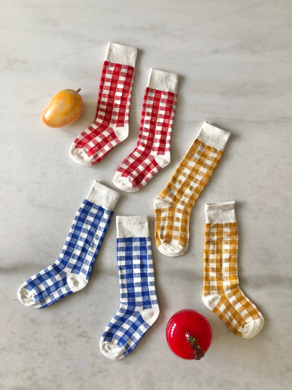 Six children's socks arranged on a light marble surface in a flat lay. Three socks are red and white gingham, three are blue and white gingham, and two are mustard yellow and white gingham.