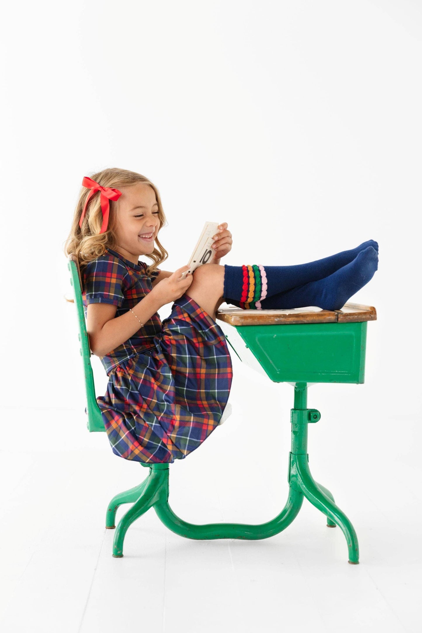 Young girl sitting on a green school desk wearing a plaid dress and blue shoes.