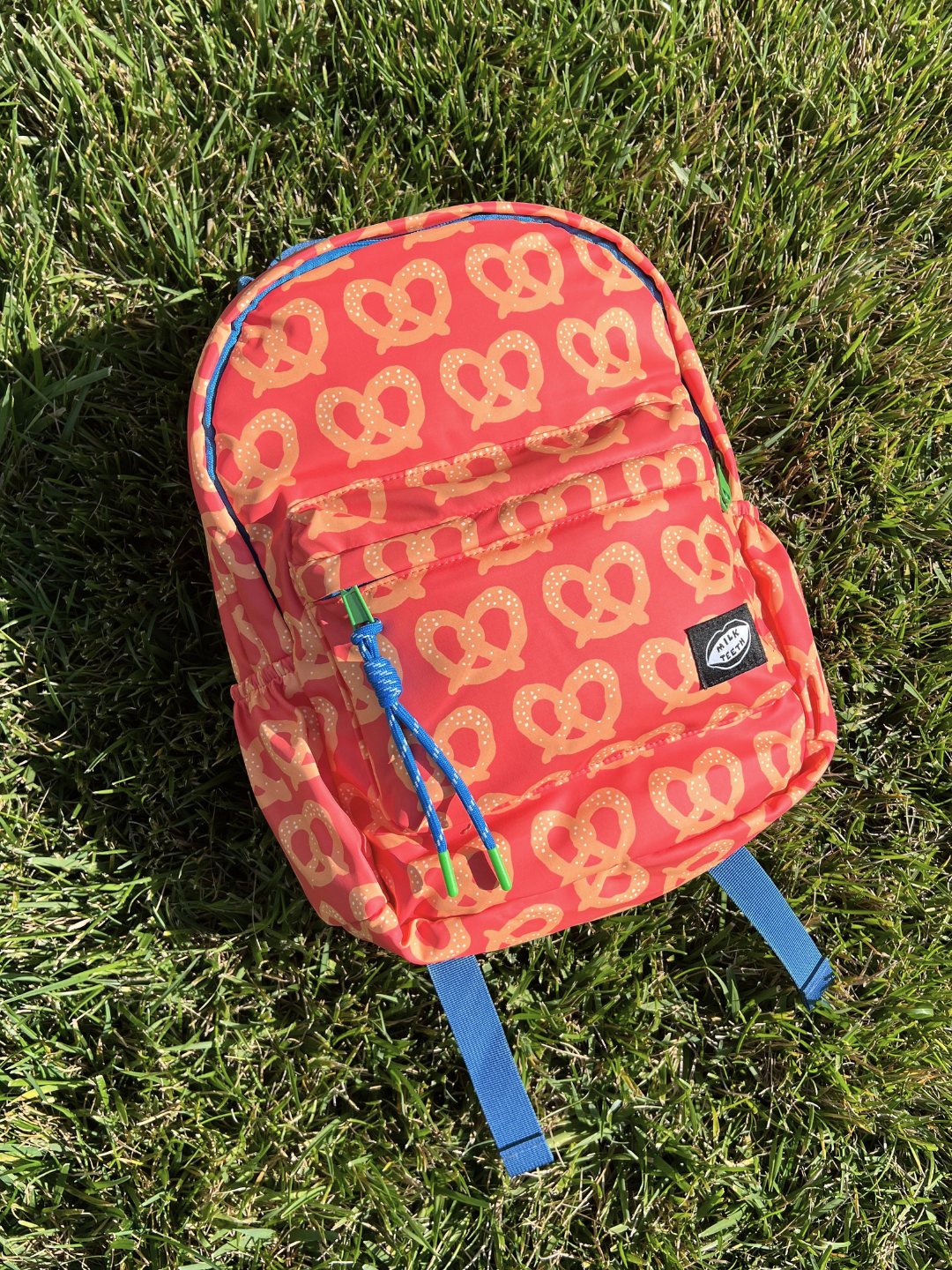 A small red backpack with an all-over tan pretzel print, resting on green grass. The backpack features blue straps and zipper pulls, and a black brand patch on the front pocket.