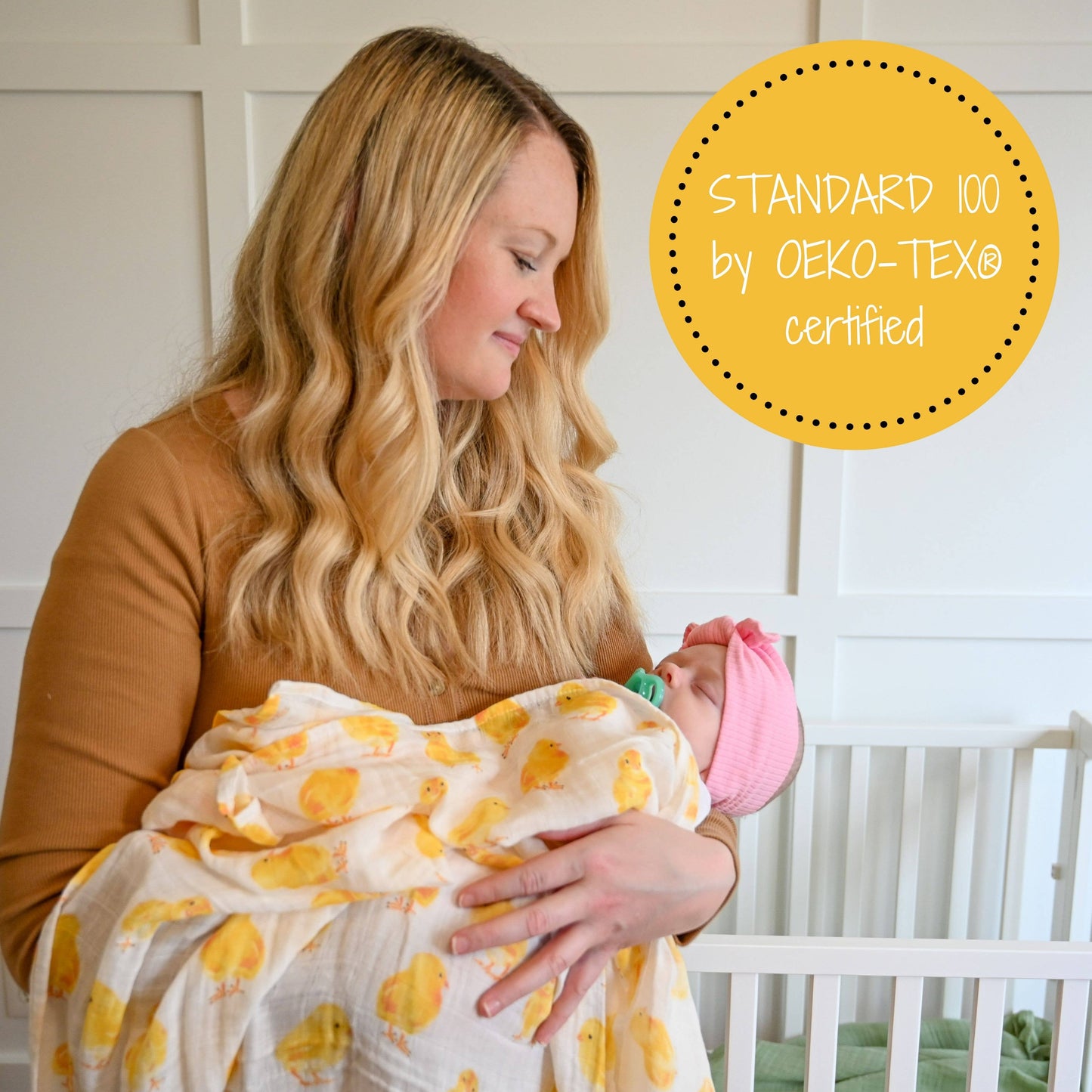 Woman holding a baby wrapped in a yellow and white patterned blanket with a crib in the background.