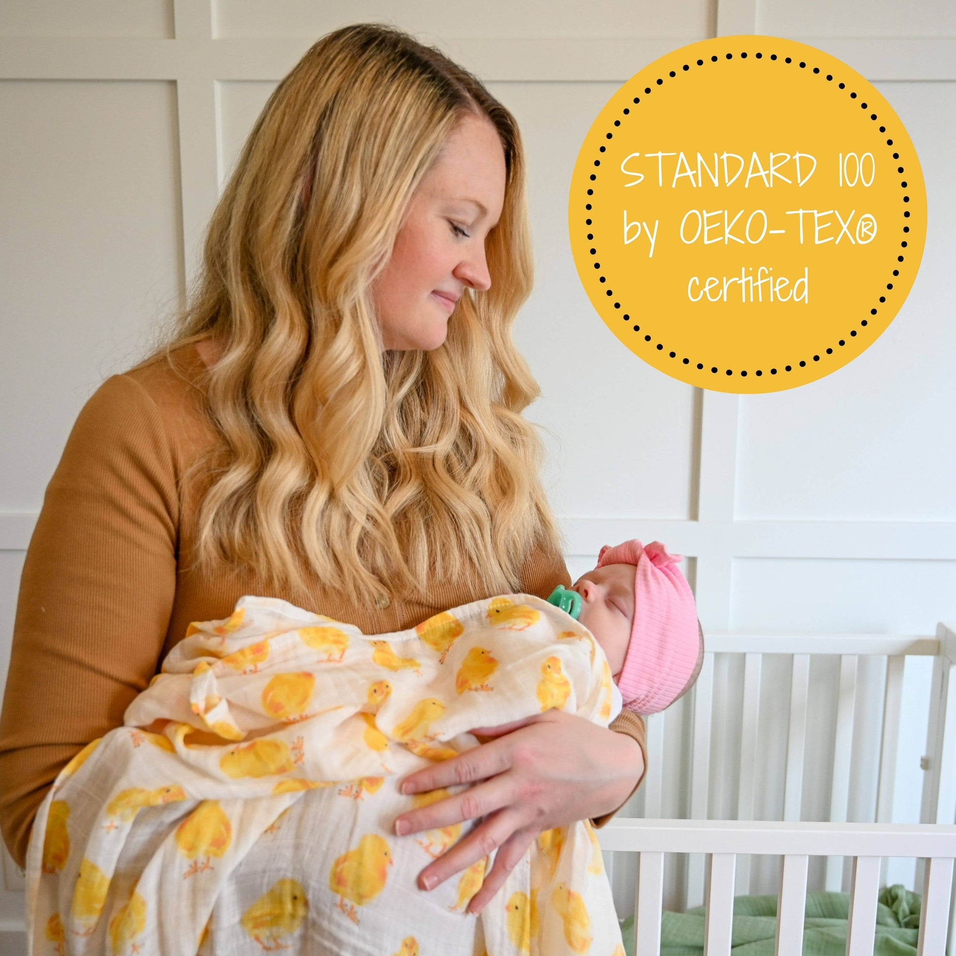 Woman holding a baby wrapped in a yellow and white patterned blanket with a crib in the background.