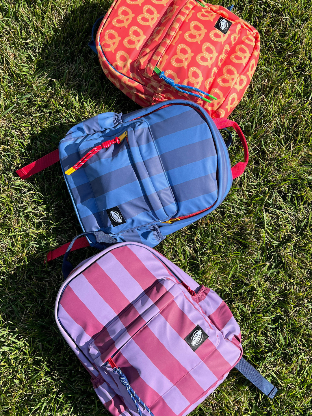 Three backpacks in different color patterns (red/orange with yellow pretzel pattern, blue and navy stripes, and pink and purple stripes) arranged diagonally on green grass.