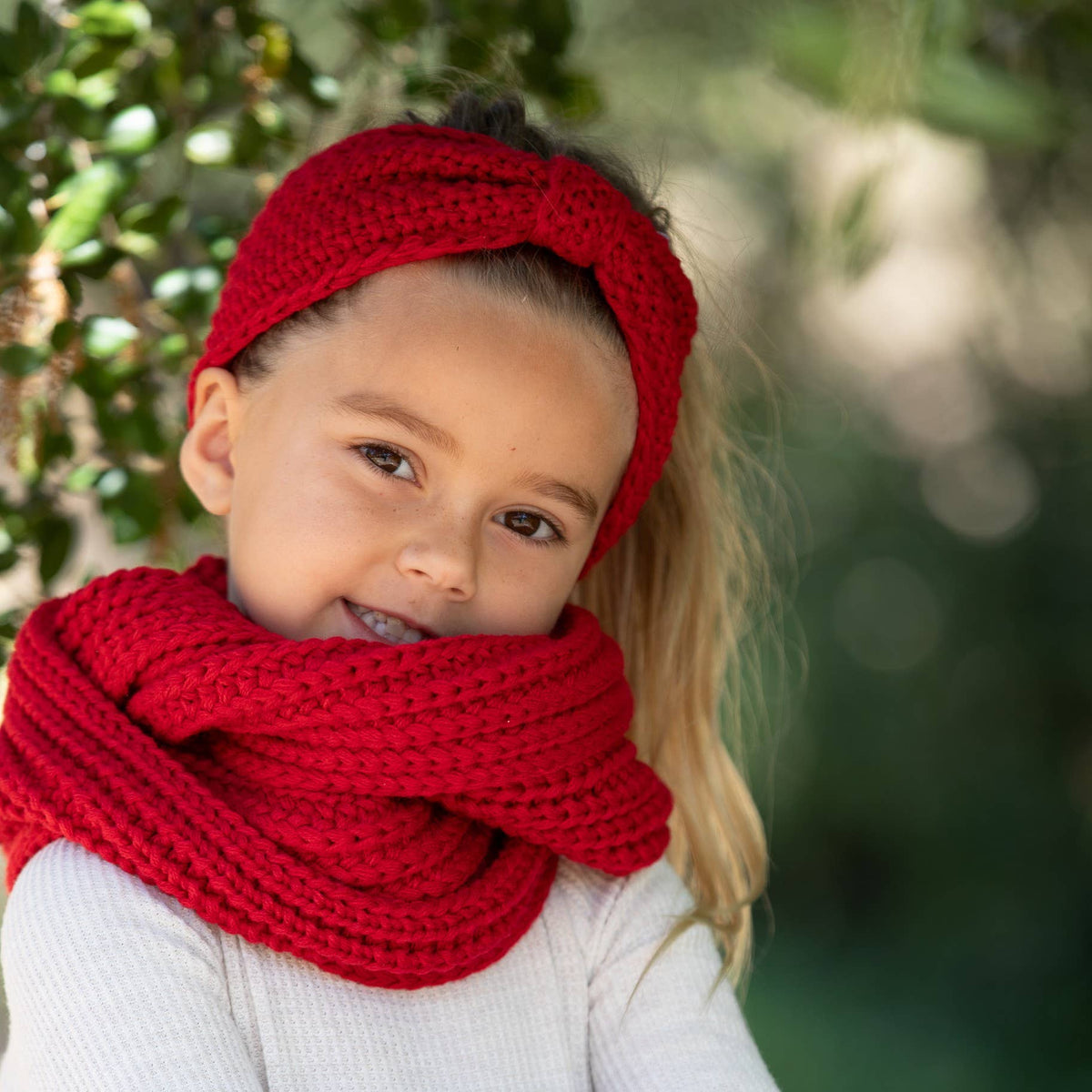 A young girl wearing a chunky red knit headband and matching red knit scarf, photographed outdoors with a soft green bokeh background.