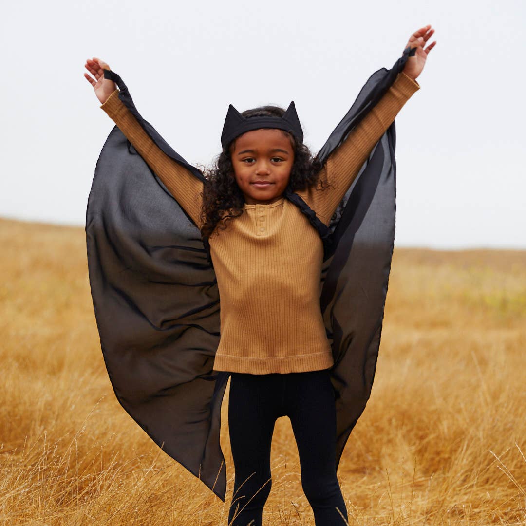 A young girl wearing a black bat cape with attached wings and a matching black bat headband, posing with her arms raised in a dry grass field.