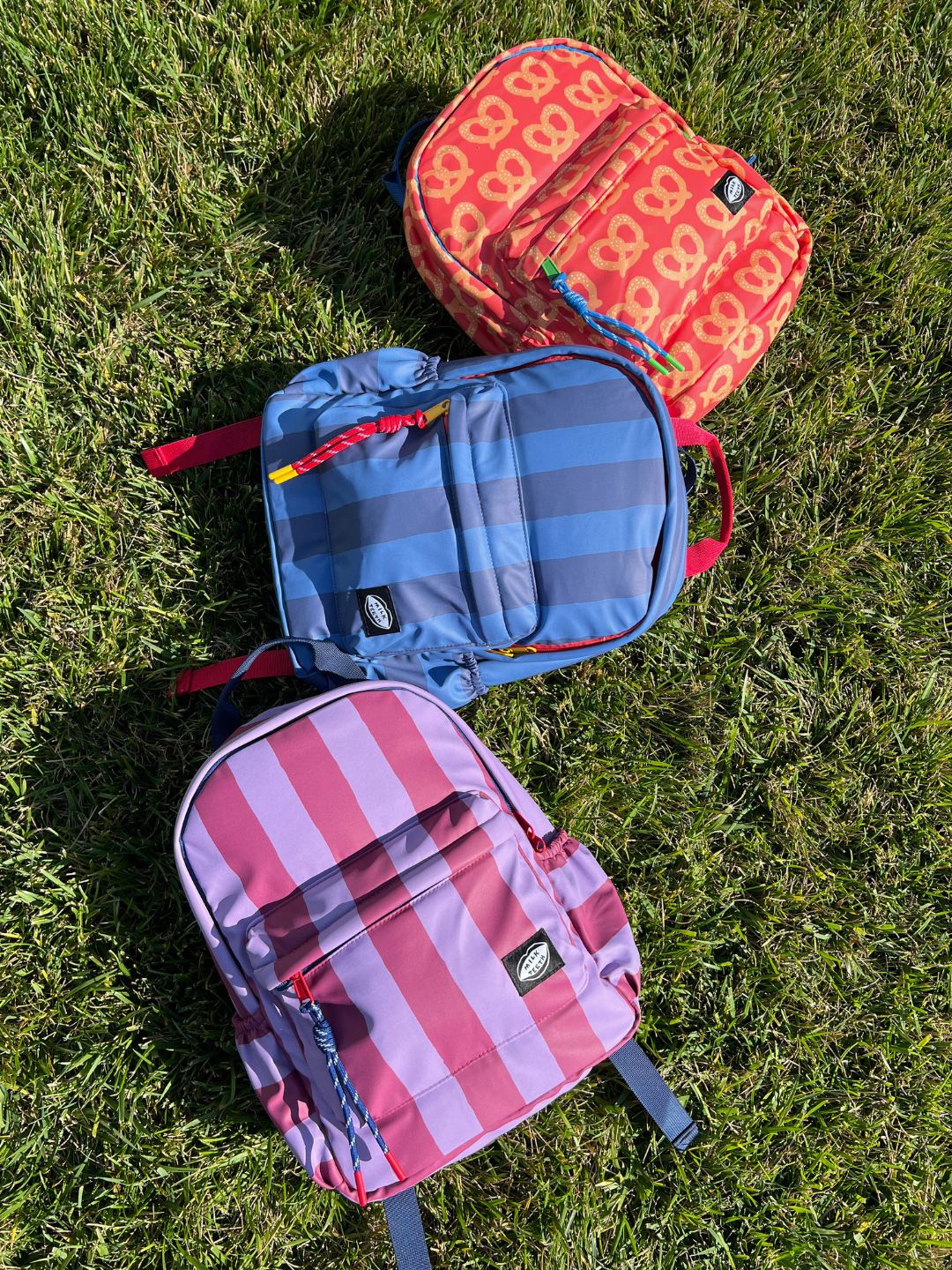 Three colorful backpacks are arranged diagonally on a green grass lawn. The top backpack is orange with a white pretzel pattern, the middle is blue with gray stripes, and the bottom is pink and purple with vertical stripes.