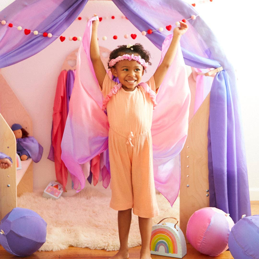 Child in a peach dress with fairy wings standing in a playroom with colorful decorations.