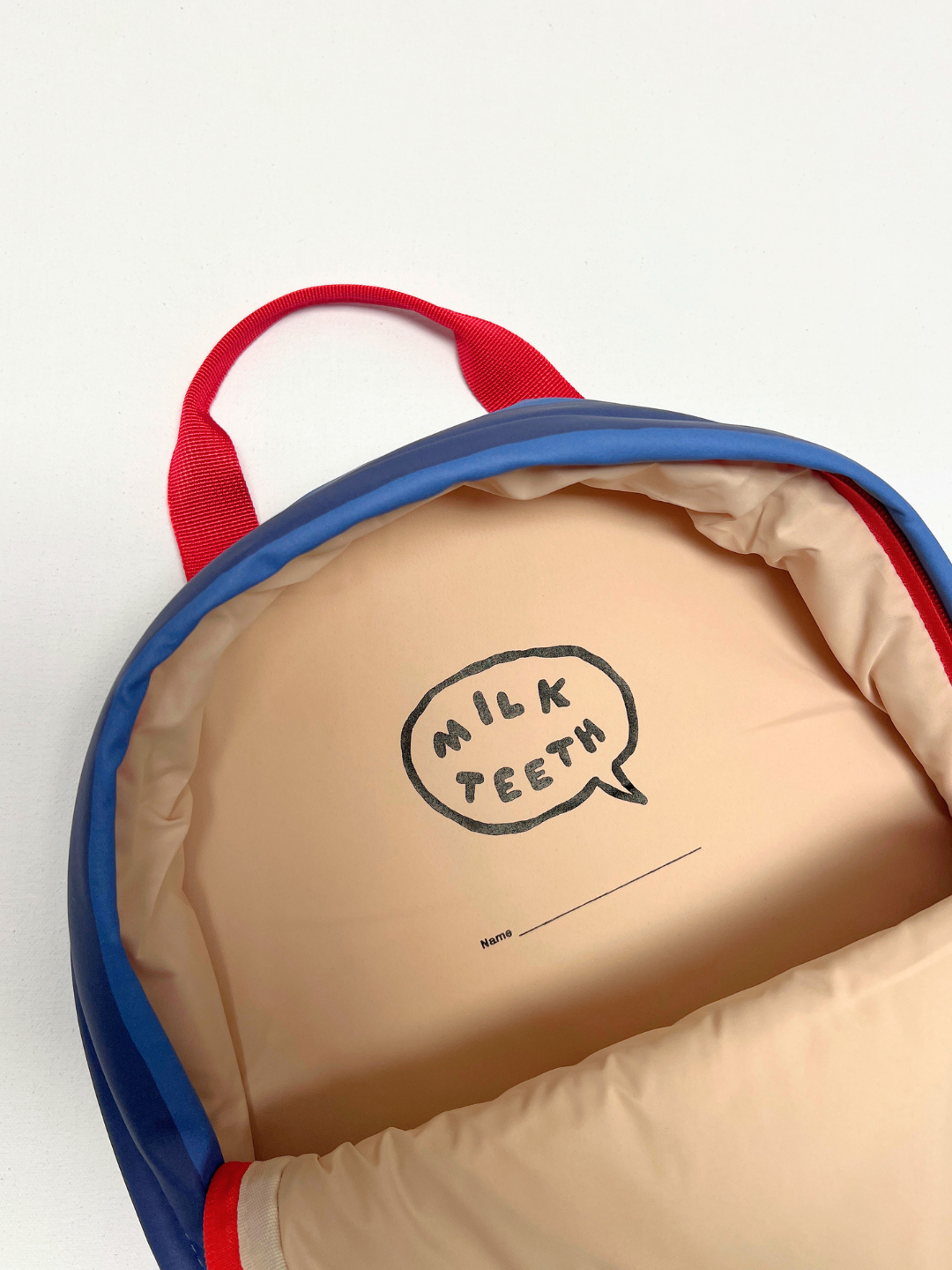 An open blue and red backpack showing a beige interior with a hand-drawn speech bubble that reads 'MILK TEETH'.