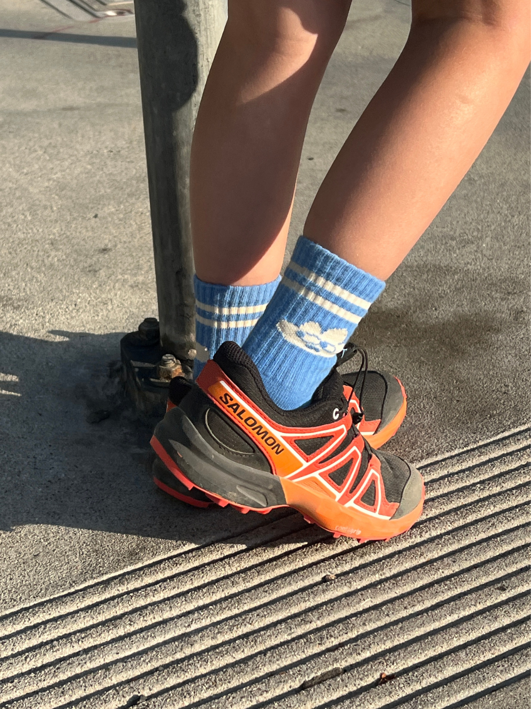 Person wearing blue socks with white stripes and black and orange Salomon shoes on a concrete surface.