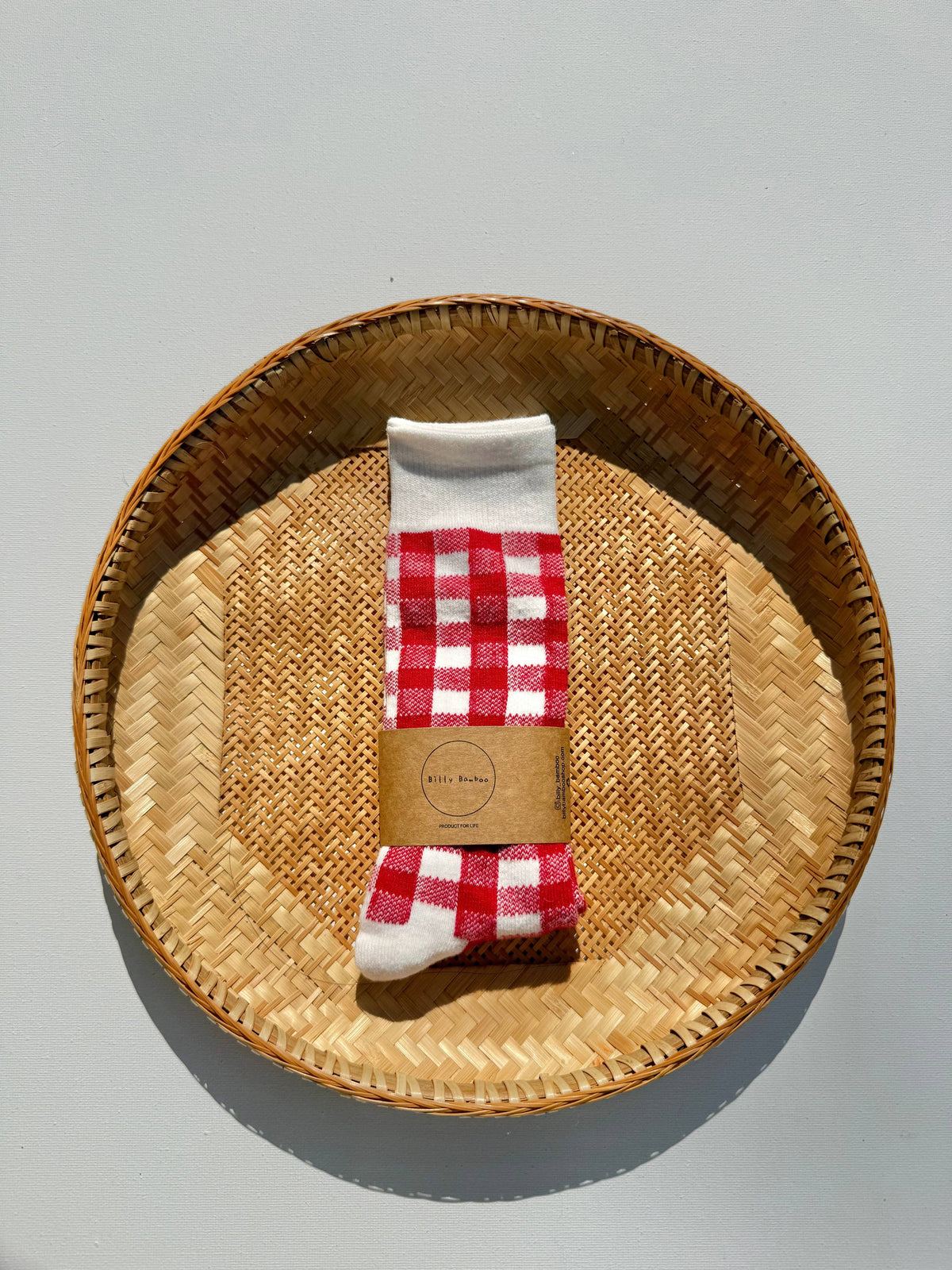 A pair of white socks with a red and white gingham check pattern, wrapped in a brown paper band, displayed on a round woven straw tray against a light gray background.