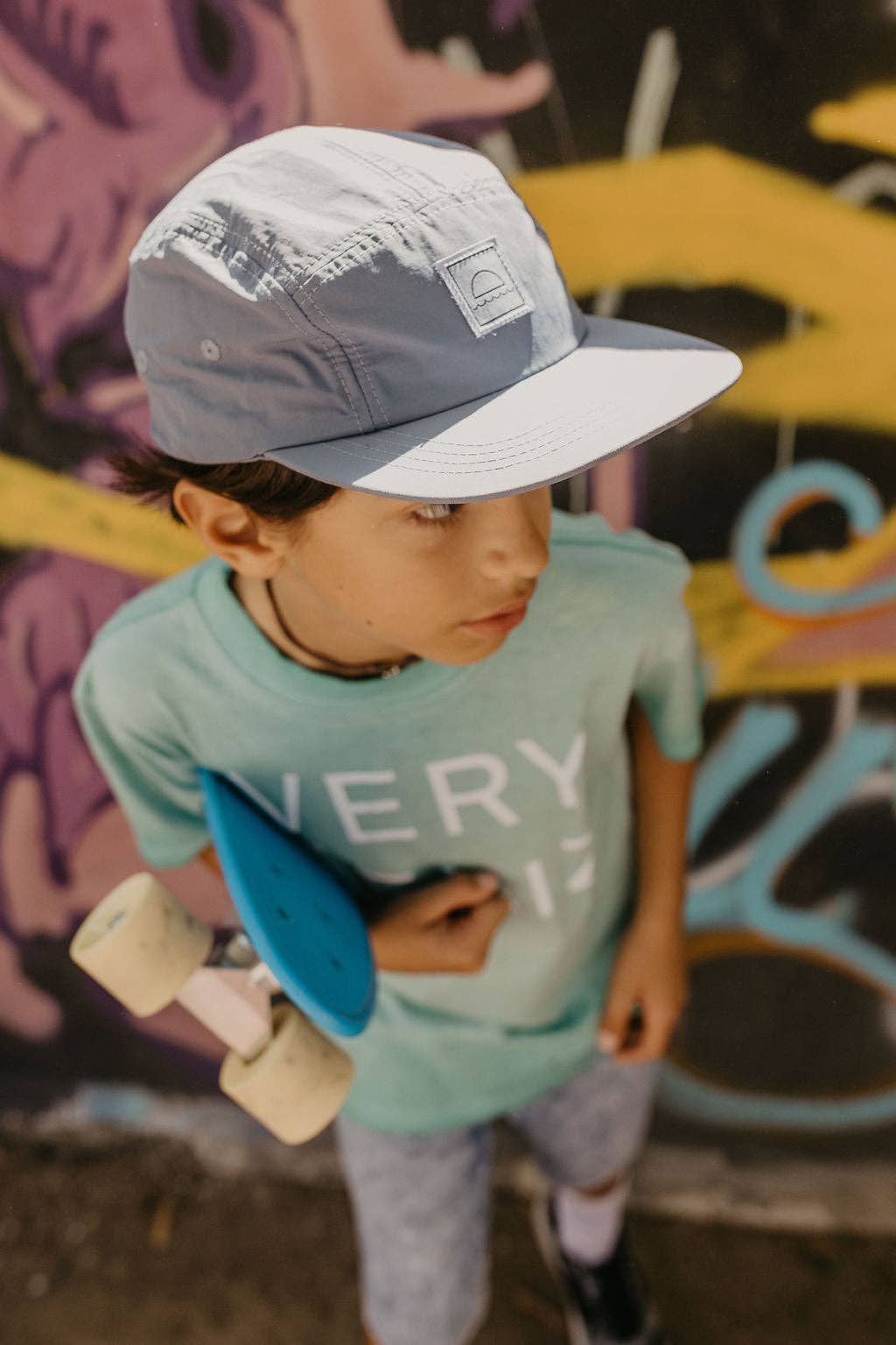 Child wearing a cap and holding a skateboard in front of a colorful graffiti wall
