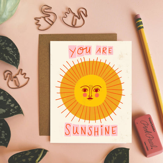 Card with sun illustration and 'You are sunshine' text on a pink background with plants and stationery items.