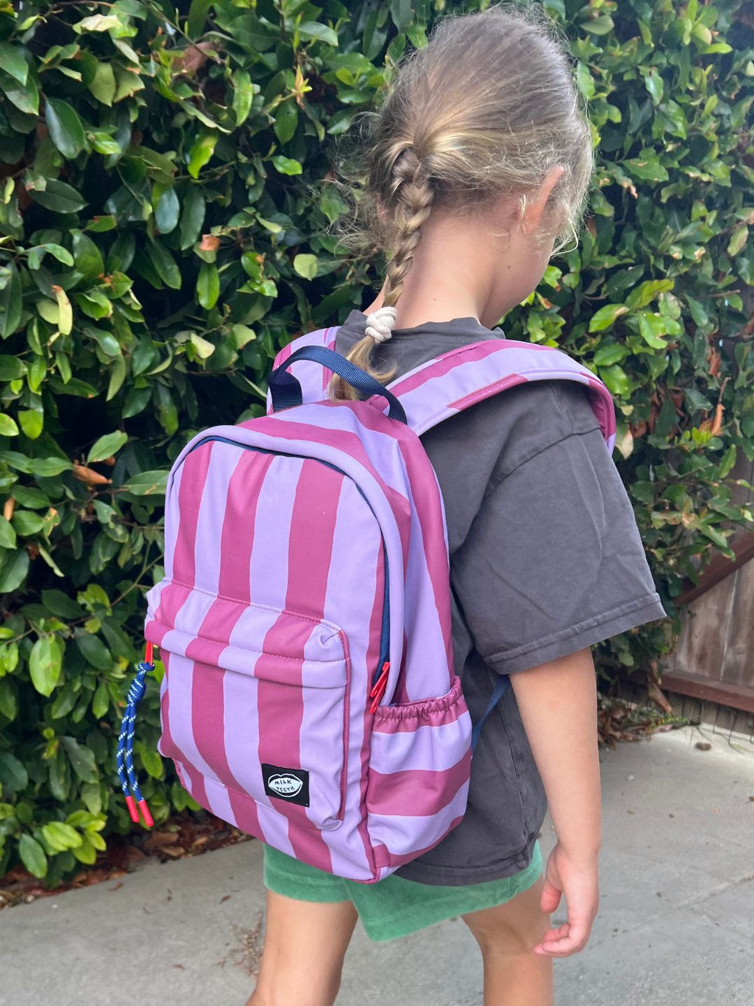 A child wearing a pink and lavender striped backpack, standing outdoors in front of a green hedge.