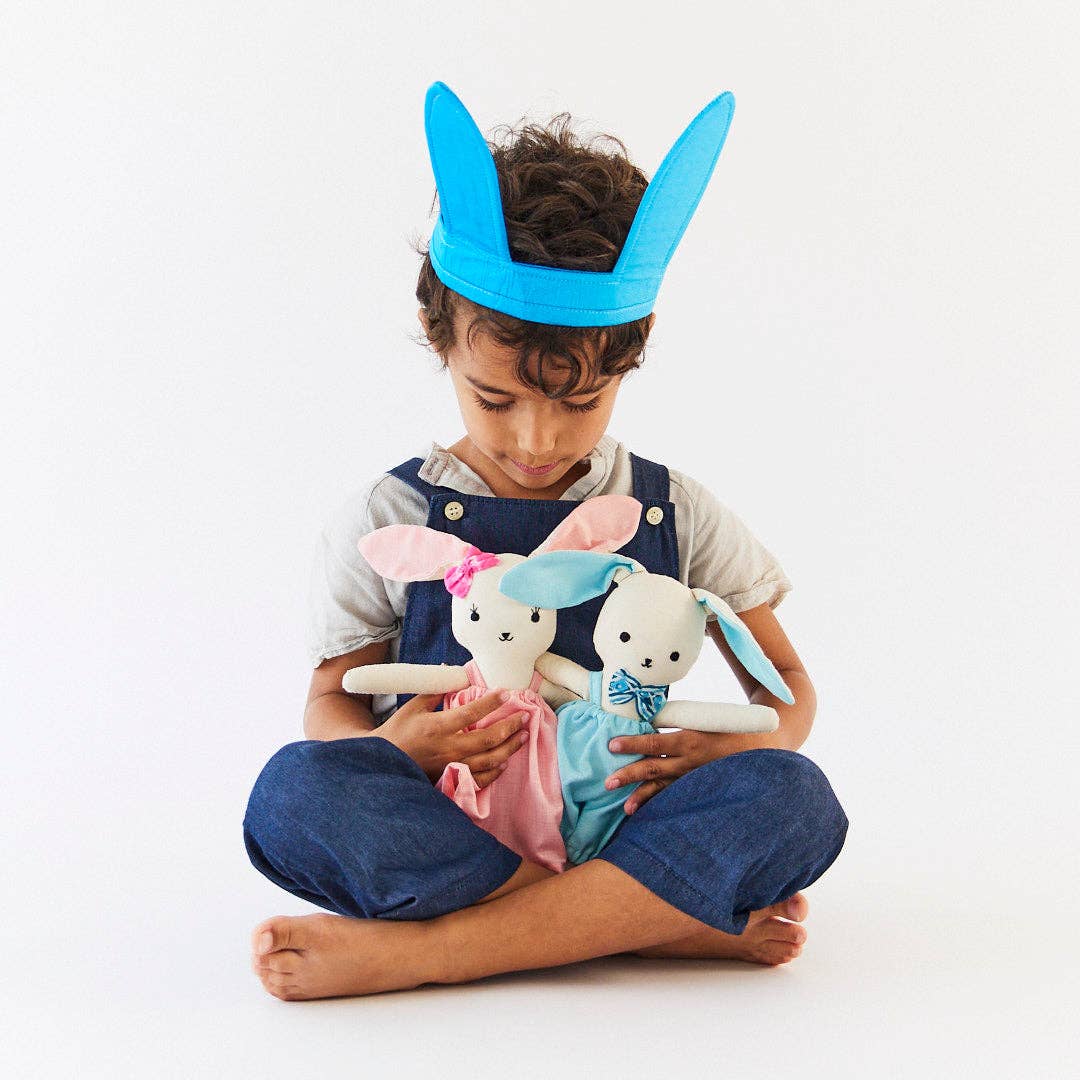 Child wearing a blue bunny headband holding two plush toys on a white background