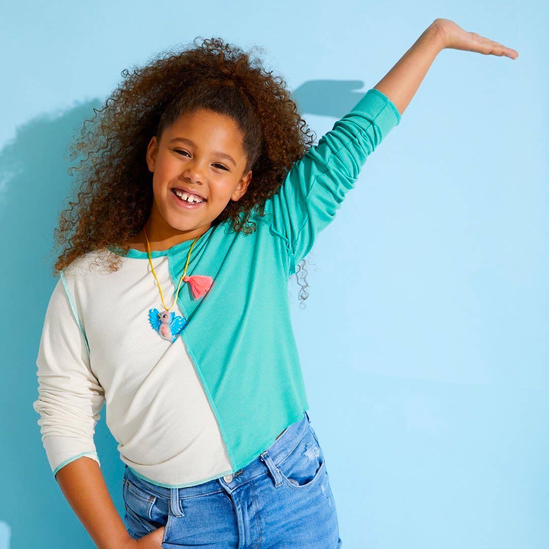 A young girl with curly brown hair smiling and posing in a color-blocked long-sleeve shirt with a teal and white split design, blue jeans, and a colorful heart necklace, set against a solid light blue background.