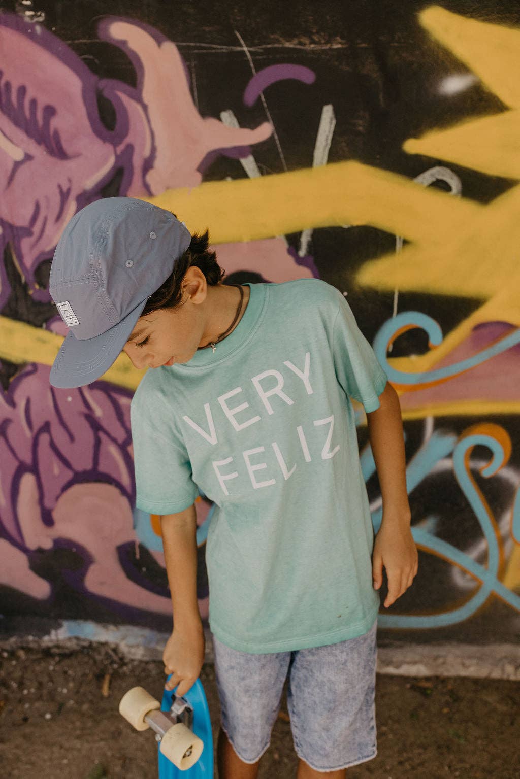 Child wearing a 'VERY FELIZ' shirt in front of a graffiti wall