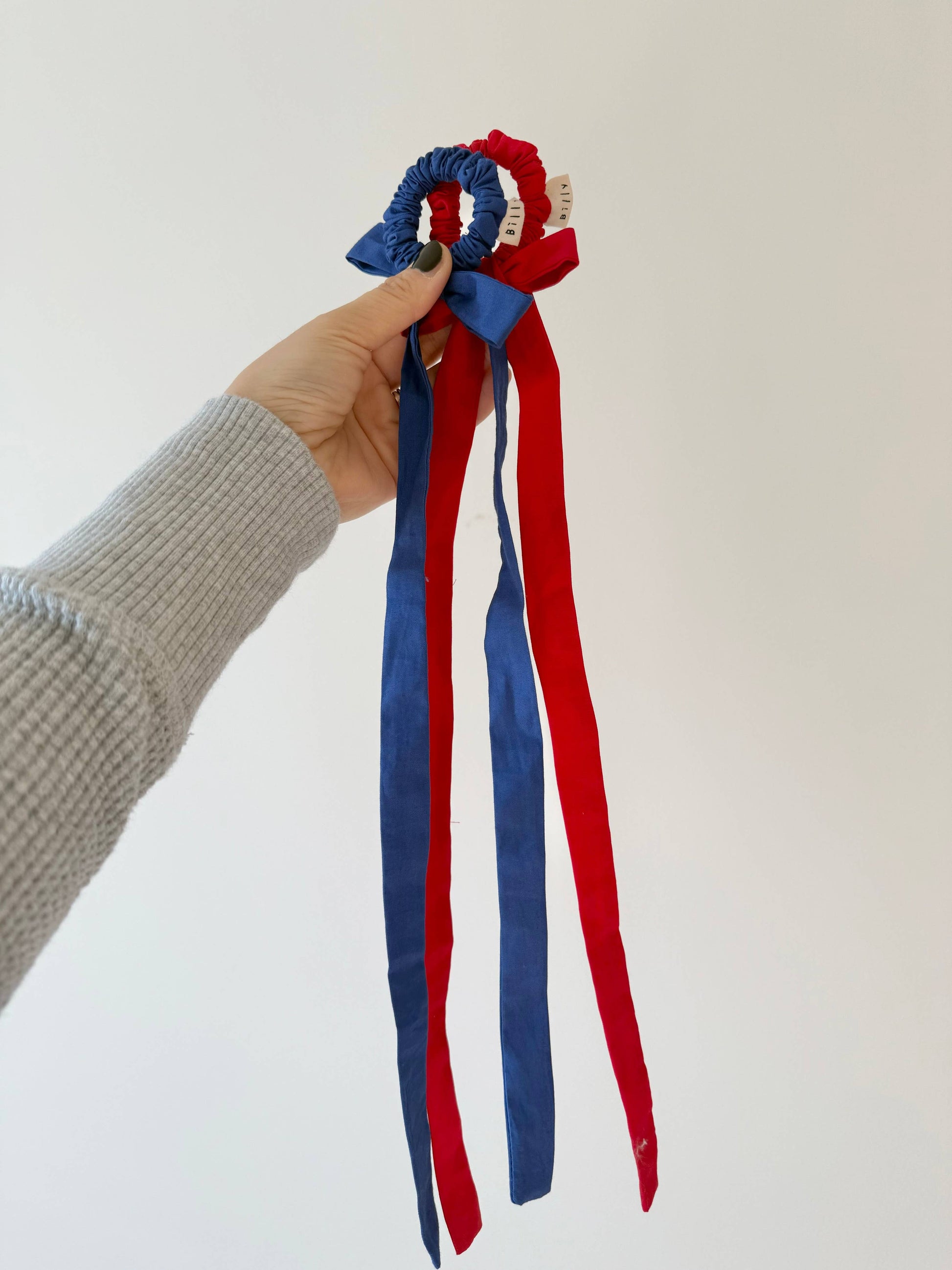 Hand holding a red and blue hair bow with long ribbons against a white background