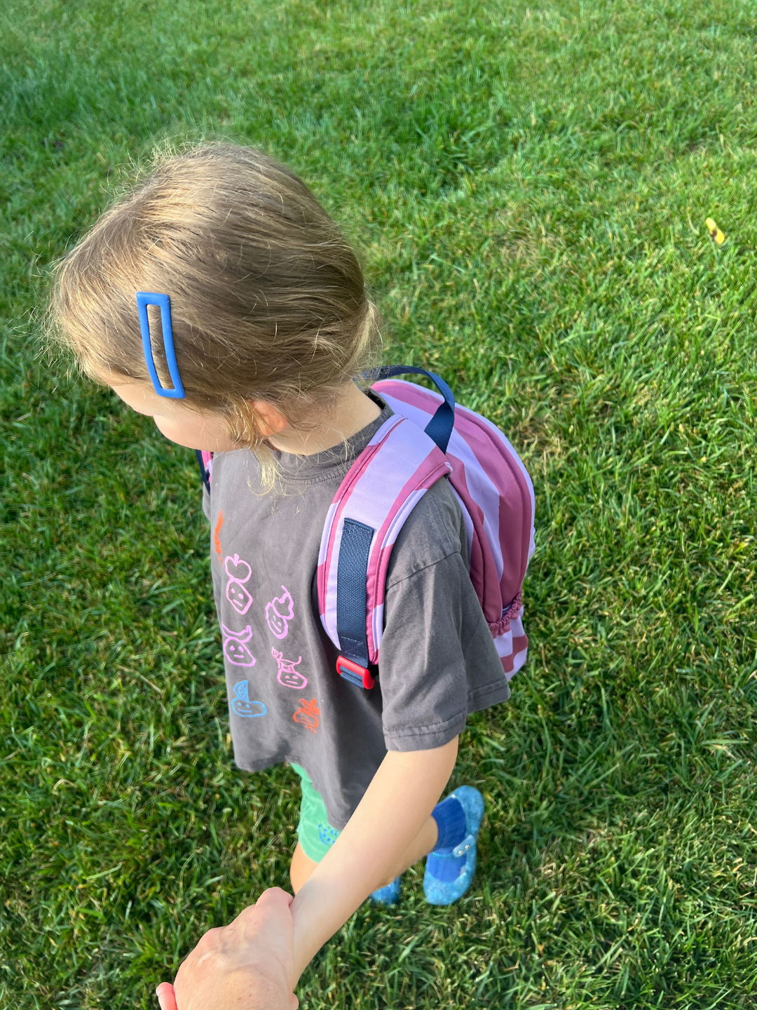 A young child walking outdoors on green grass, wearing a gray graphic t-shirt, blue sneakers, and a pink striped backpack.