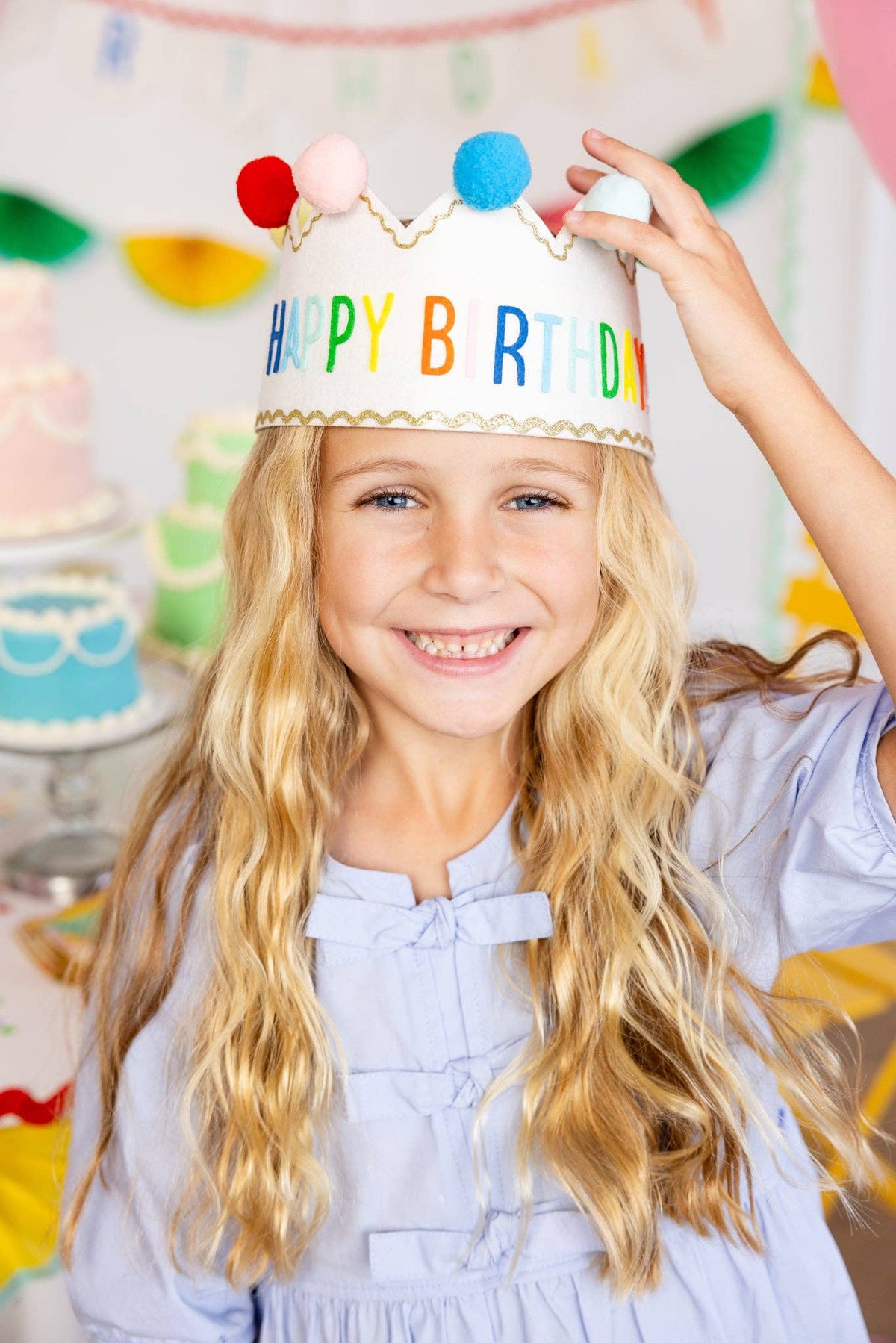 Child wearing a 'Happy Birthday' crown with colorful pom-poms, standing in front of a festive background.