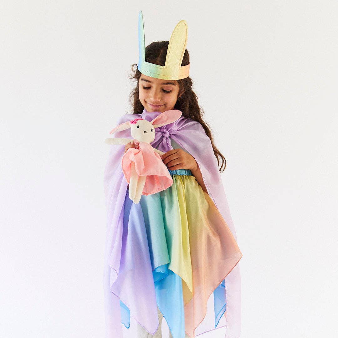 Child wearing a colorful unicorn costume with a headband and cape, holding a plush toy on a white background.