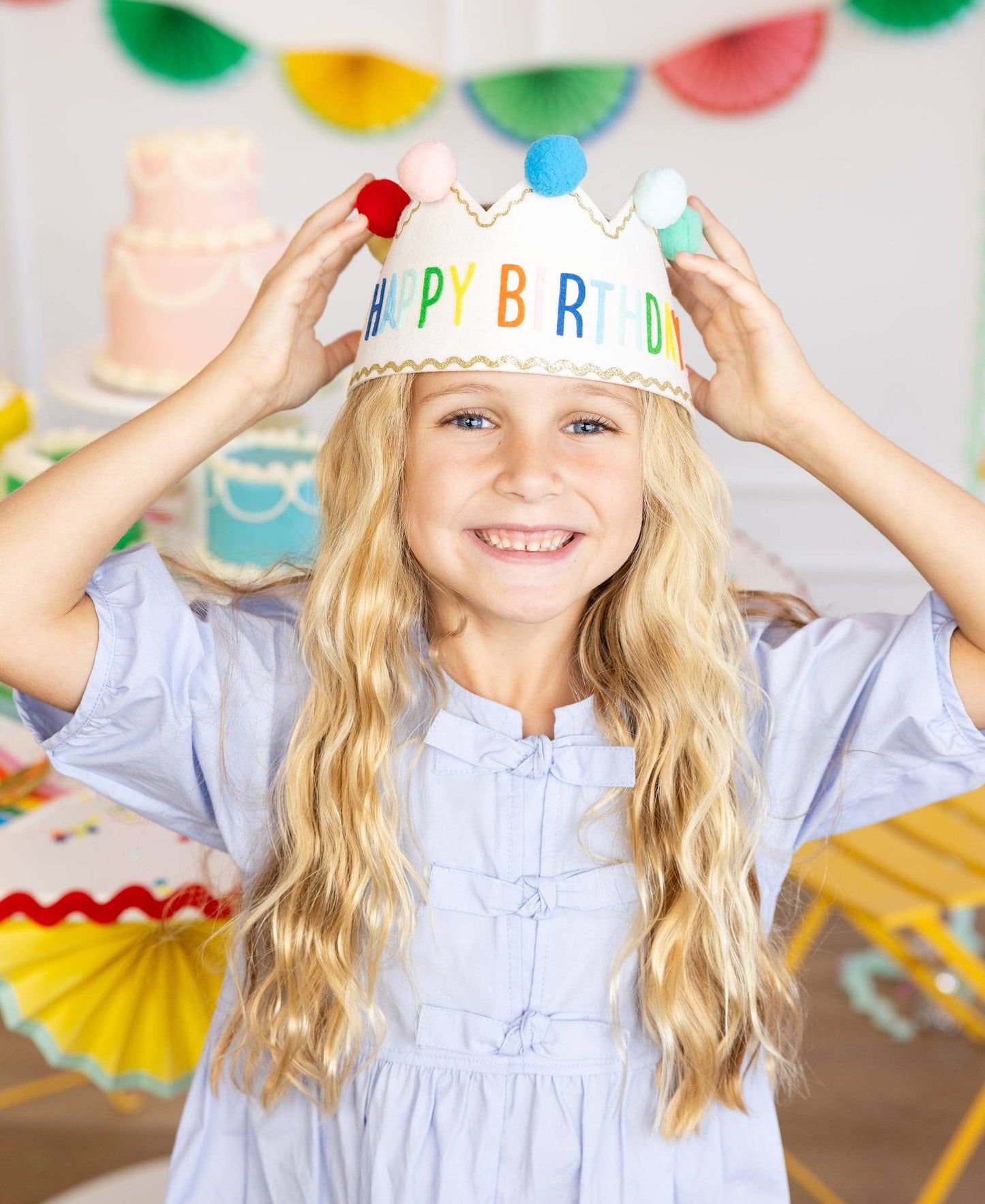 Child wearing a 'Happy Birthday' crown with colorful letters, standing in front of a birthday cake and decorations.