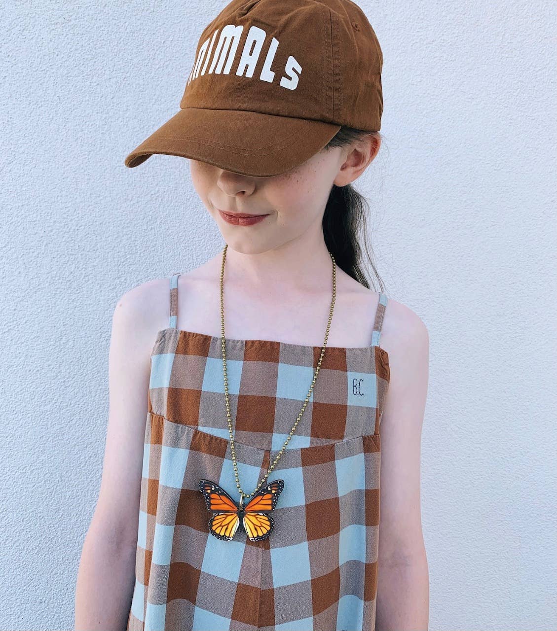 Child wearing a brown cap with 'ANIMALS' text, plaid dress, and butterfly necklace against a light blue background