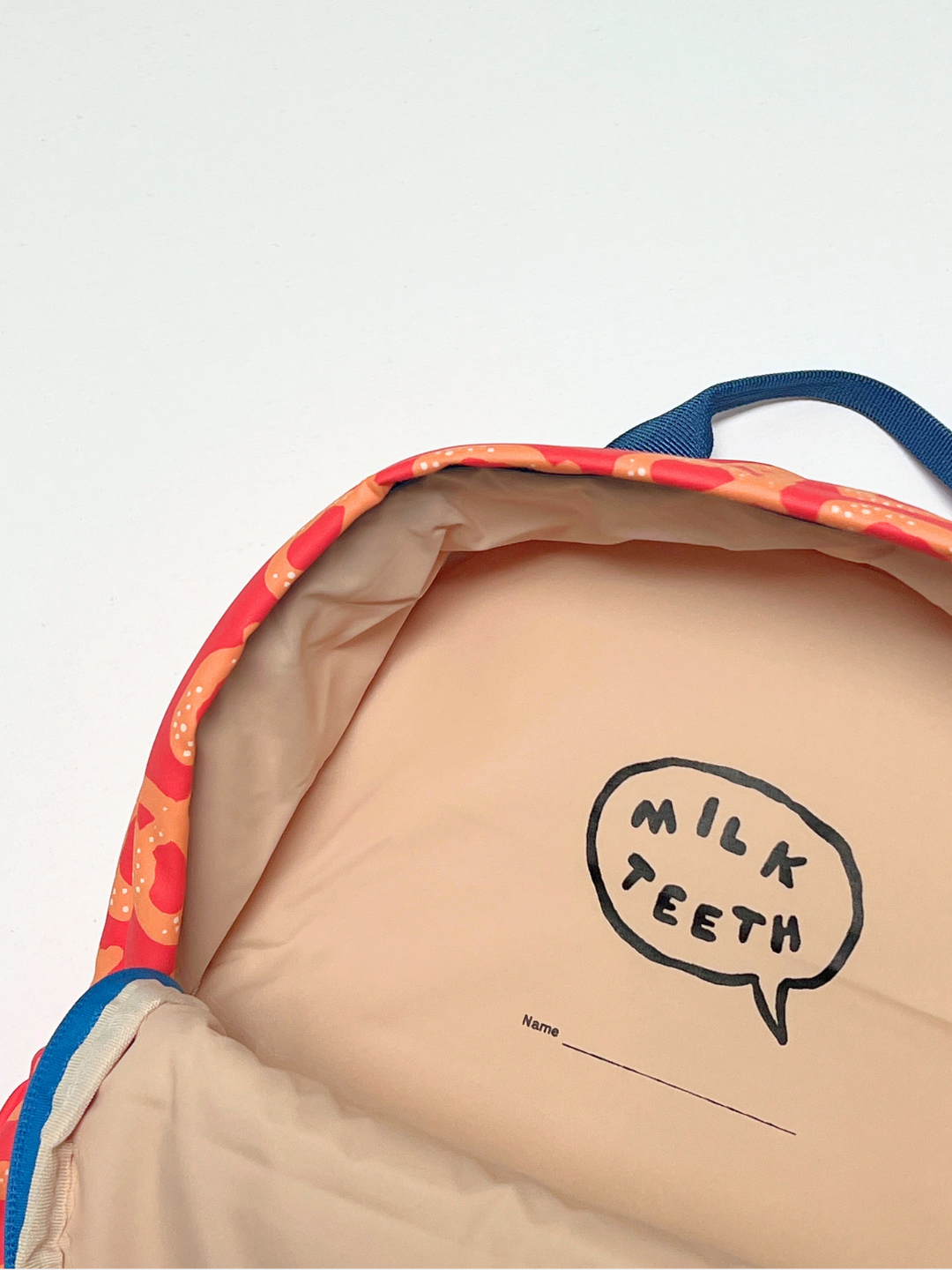 Close-up of the inside of a backpack showing a beige interior with a black speech-bubble logo that reads 'MILK TEETH' and a colorful patterned fabric lining with blue trim.