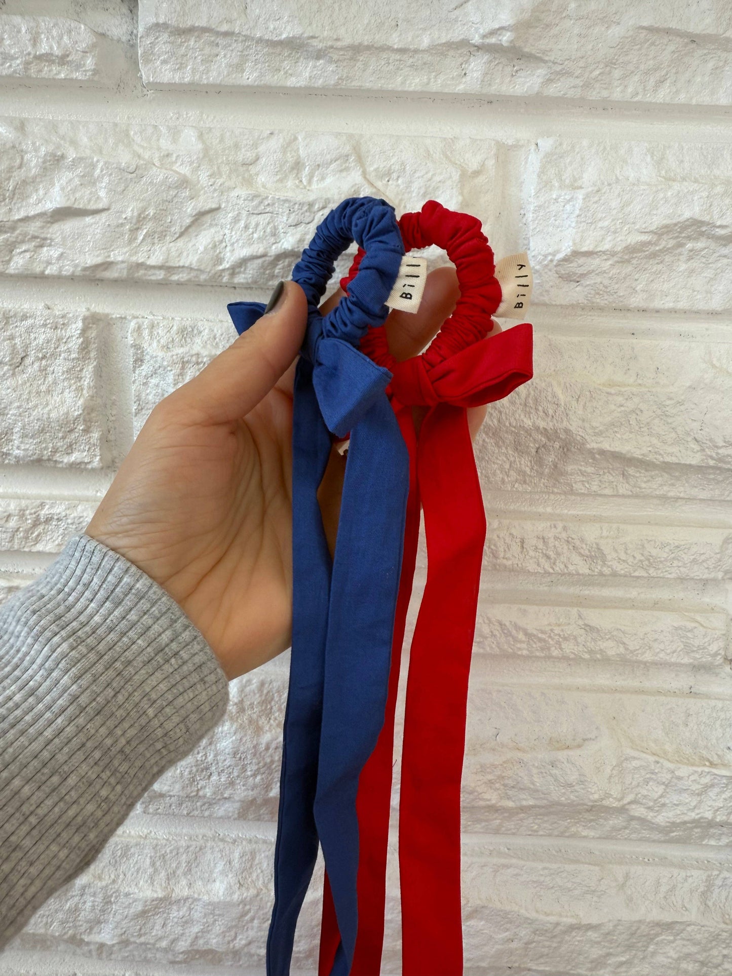 Hand holding a blue and a red scrunchie against a light stone wall background